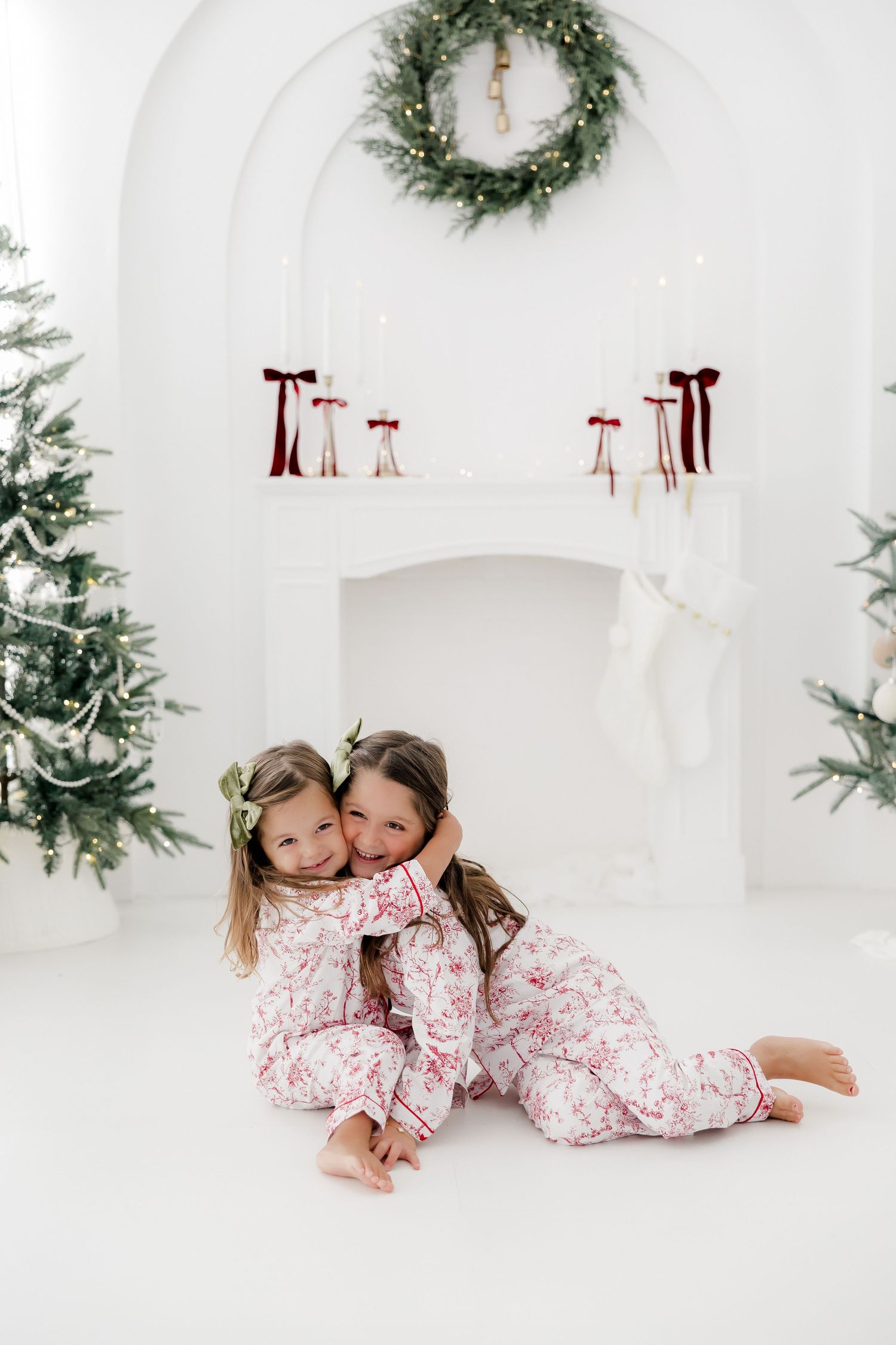 Two girls in matching floral pajamas hug near a white fireplace with Christmas wreath and tree.