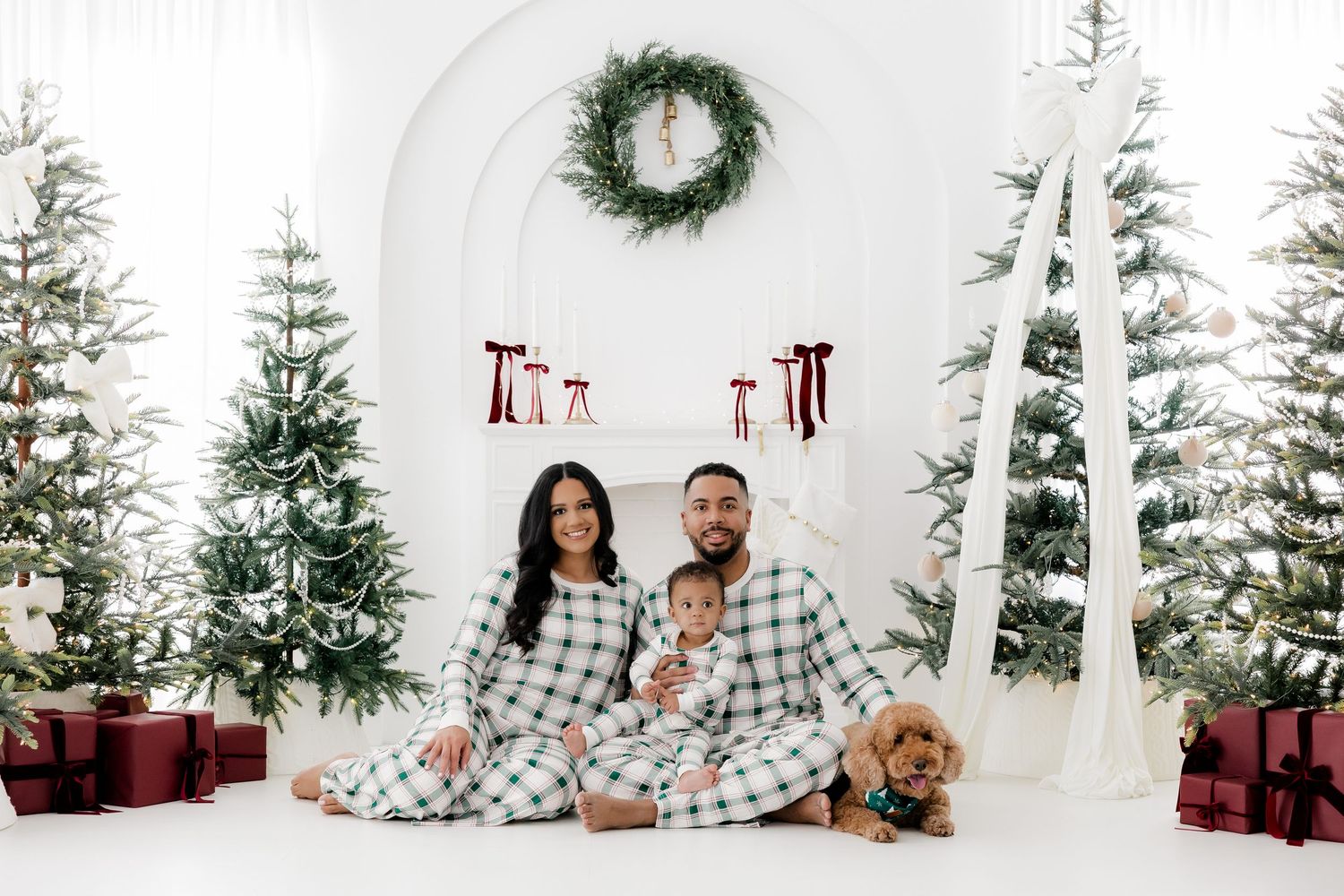 Family in matching plaid pajamas poses with a dog among Christmas trees and gifts in a festive white holiday setting.