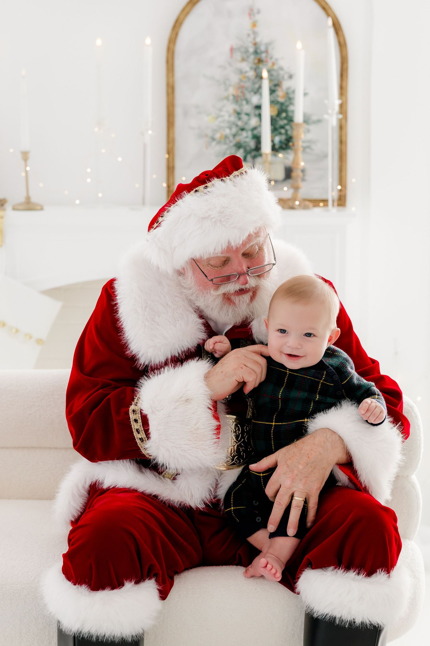 Santa Claus holds a smiling baby in a plaid outfit during a festive Christmas portrait session.