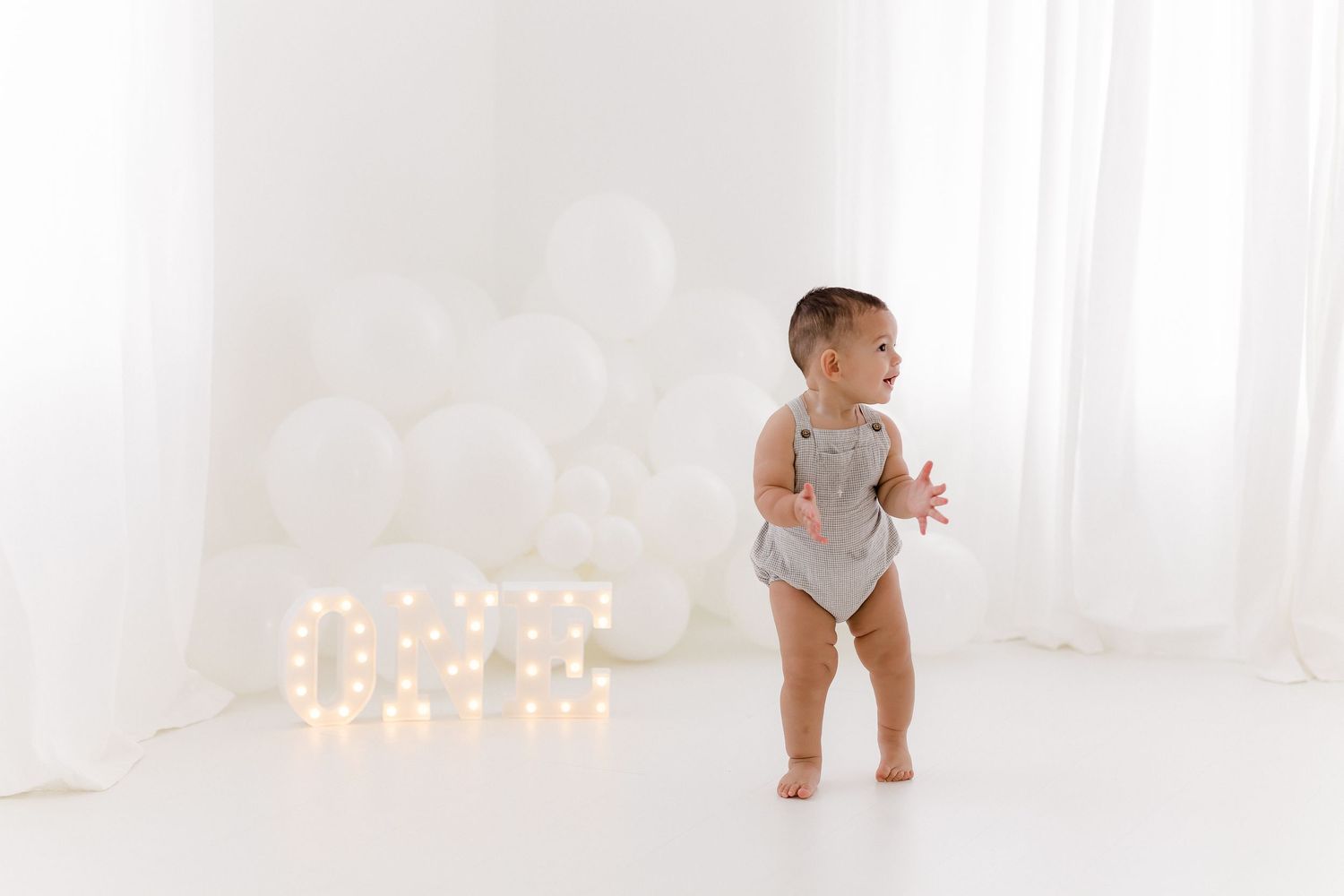 Baby in white onesie stands in front of illuminated letters against white backdrop with soft lighting.