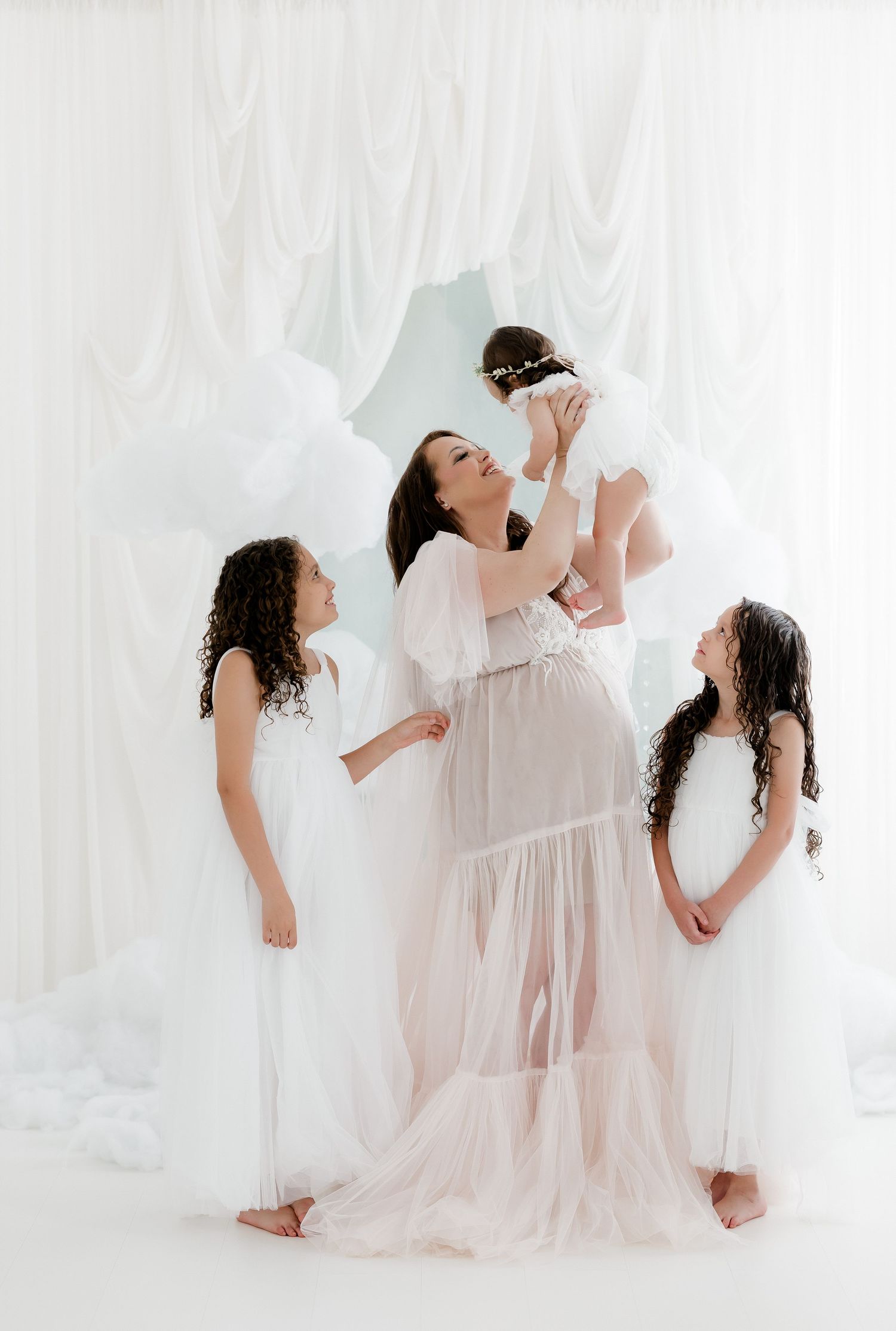 Pregnant woman in sheer white gown lifts baby girl while two daughters in white dresses stand beside her.