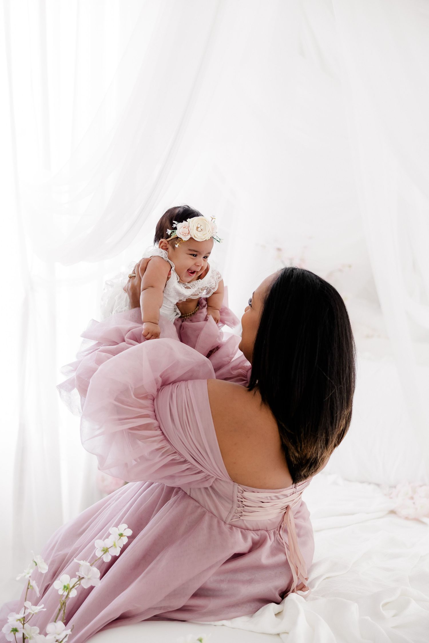 A tender moment captured in soft light as a mother in a pink dress lifts her baby wearing white against a white backdrop.