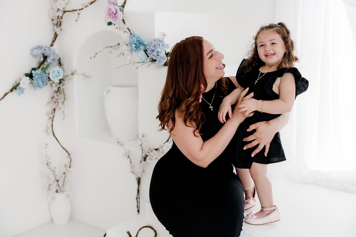 Woman in black dress holds and comforts young child against white marble backdrop in elegant studio setting.