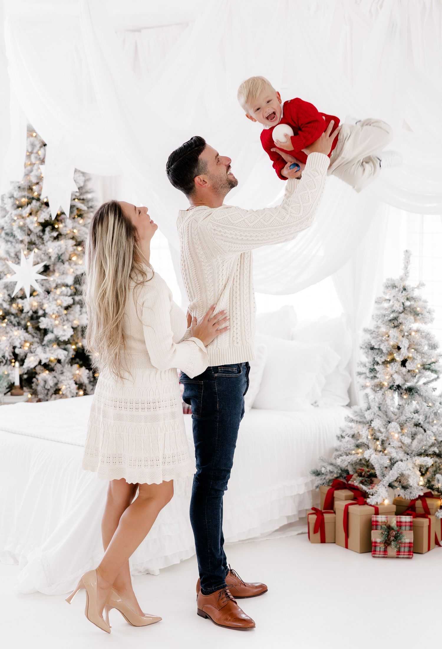 Family Christmas photo with parents lifting baby in red outfit near decorated trees and gifts.