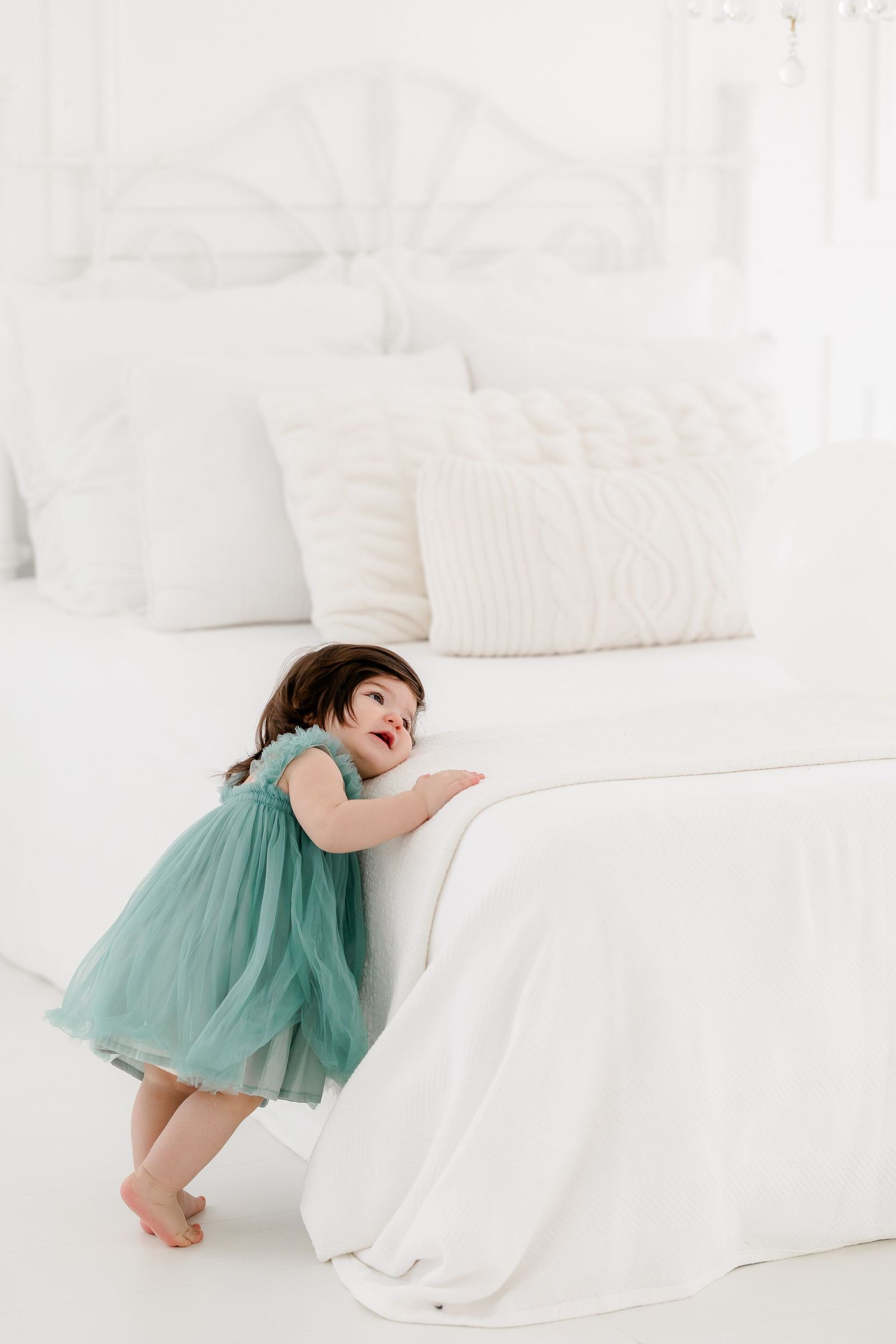 A small child in a mint green tulle dress plays and leans on a white couch in a bright minimalist setting.