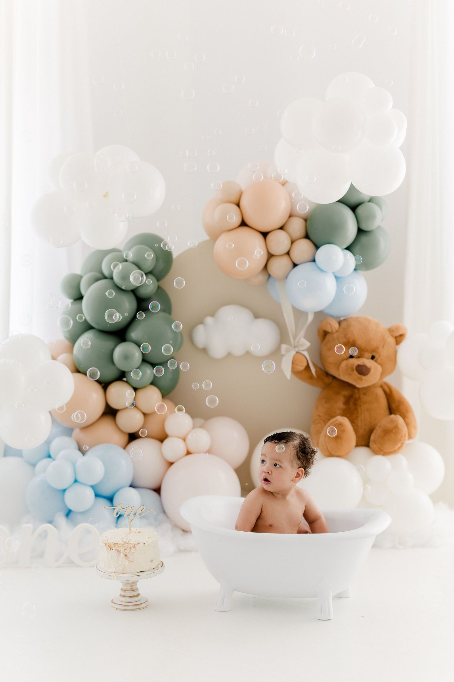 Soft pastel balloon arch installation with teddy bear and white bathtub setup for baby photography session.
