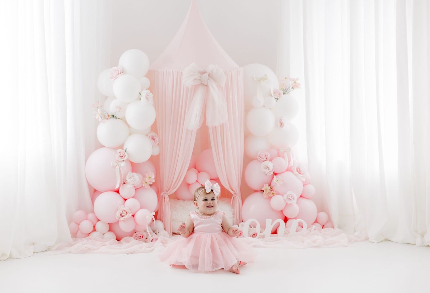 Pink and white balloon arch decorates a dreamy nursery setting with soft tulle draping and delicate decor elements.