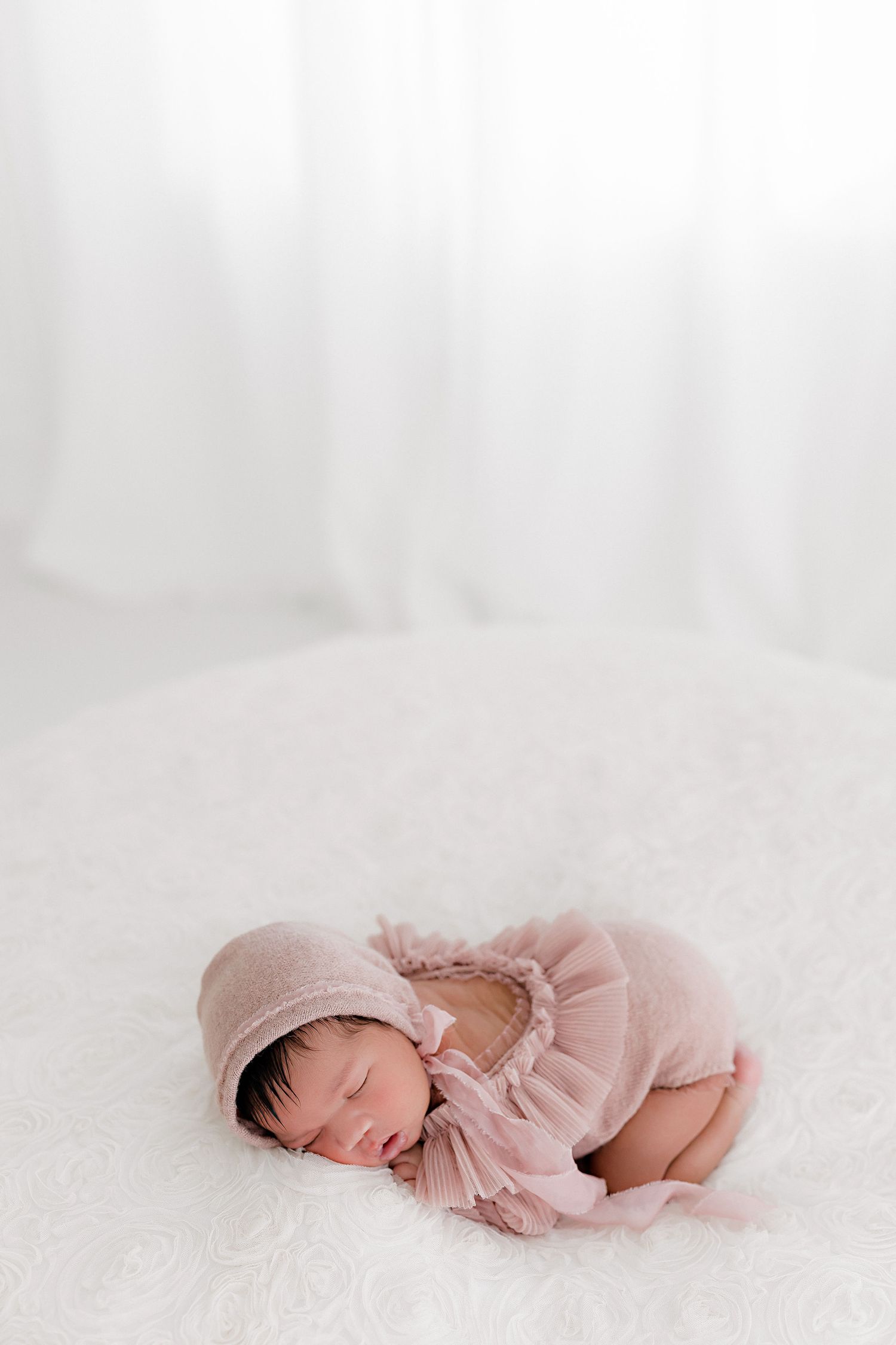 Sleeping newborn in soft pink dress laying on white textured surface.