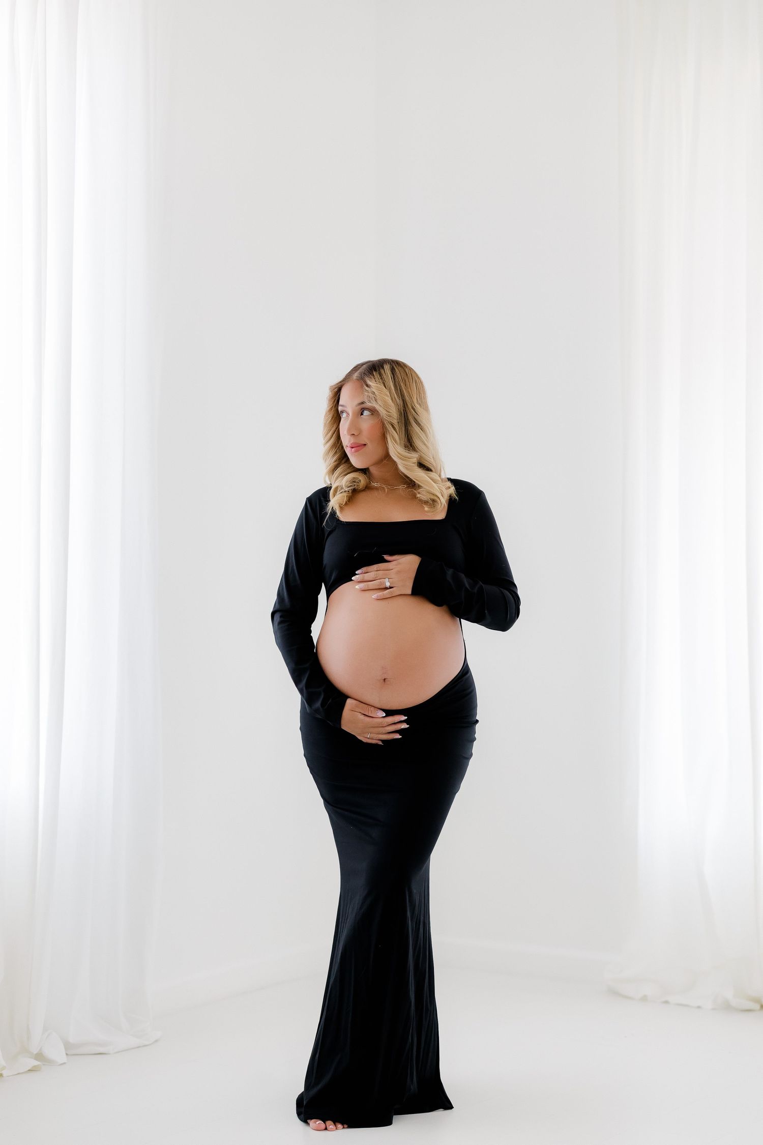 Maternity portrait in black dress against white background with soft natural lighting.