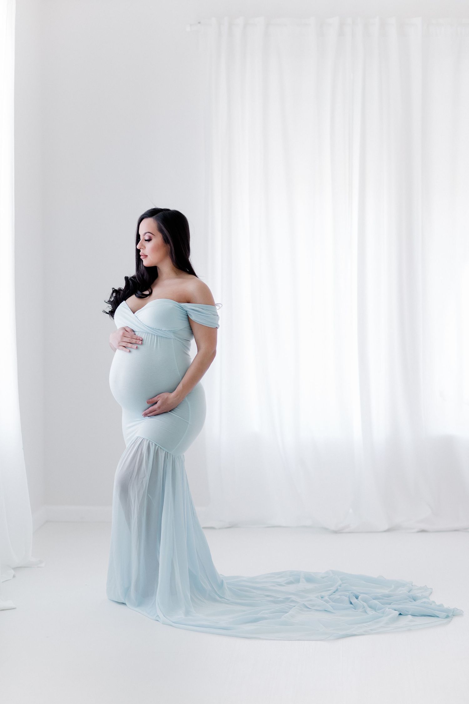 Ethereal maternity photo featuring flowy light blue off-shoulder gown with long train in white studio setting.