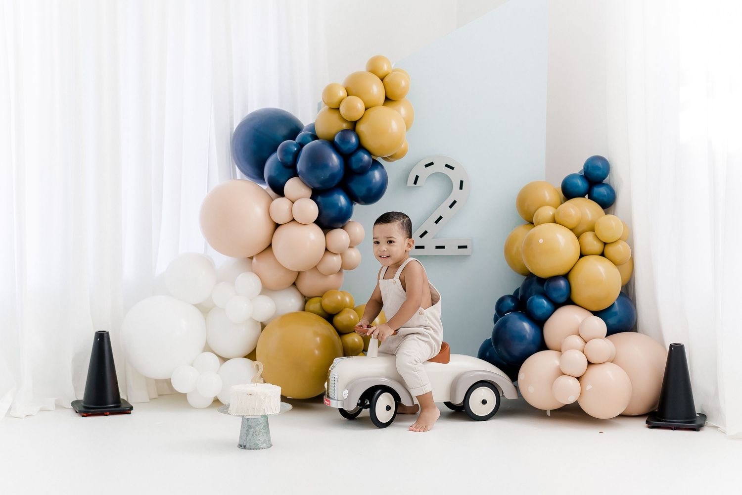 Vintage toy car setup with balloon garland decorations in navy blue, gold, and blush pink colors against white backdrop.