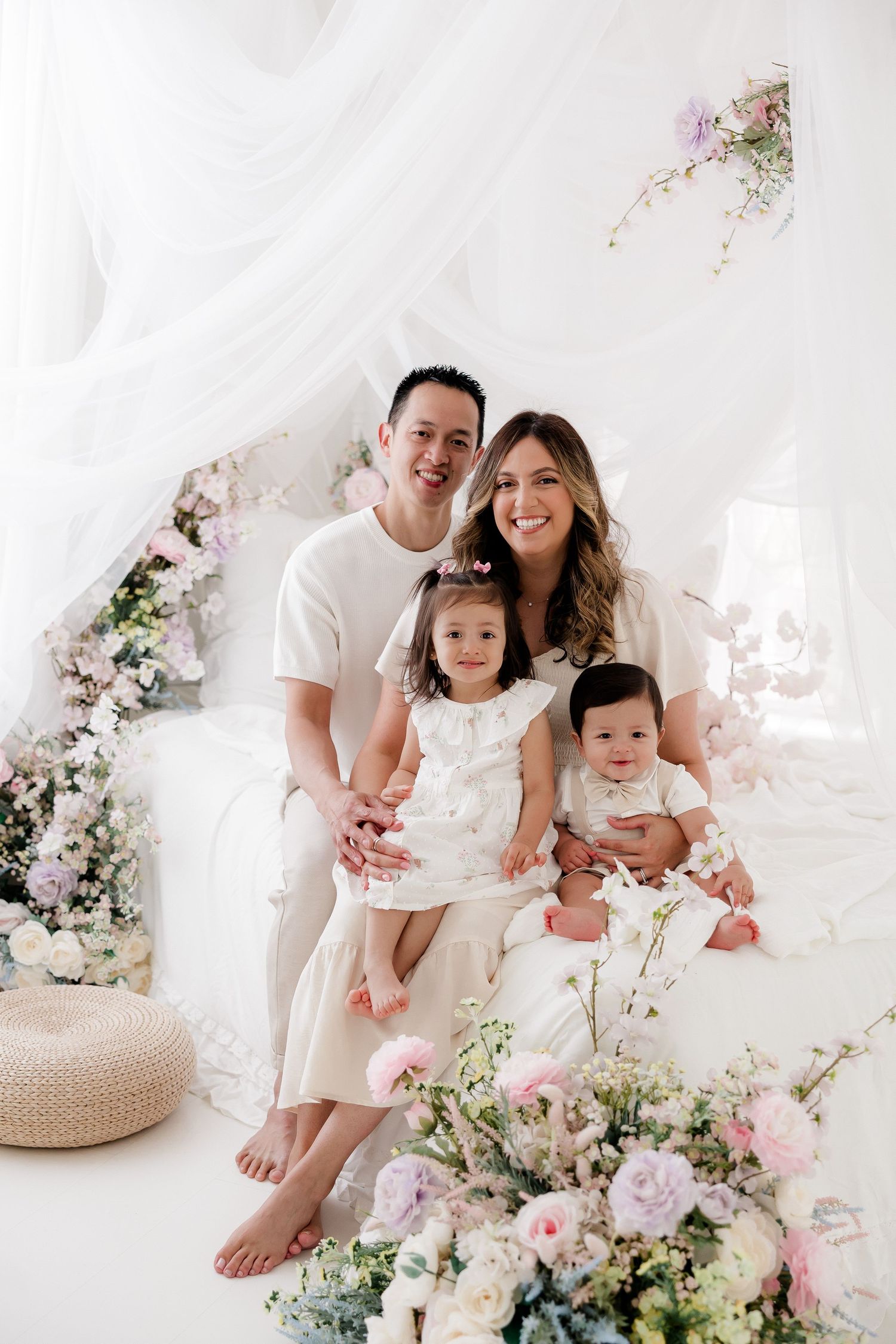A family wearing white outfits poses together surrounded by soft pink and white floral arrangements in a bright studio setting.