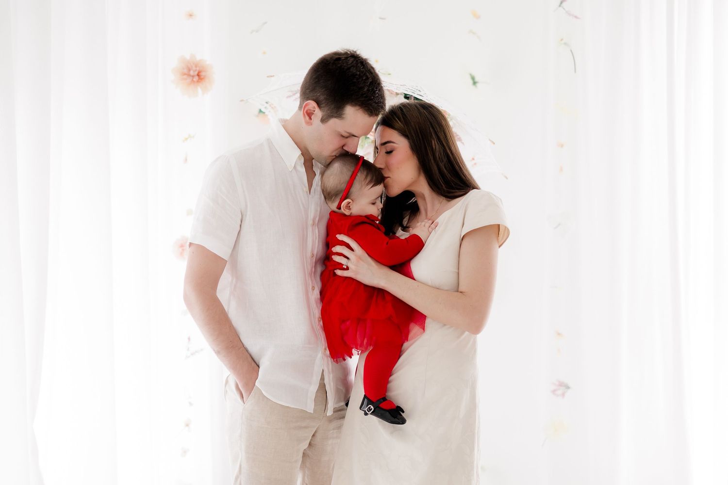 A couple in white outfits shares tender moments while holding their baby in a red outfit during a photo session.