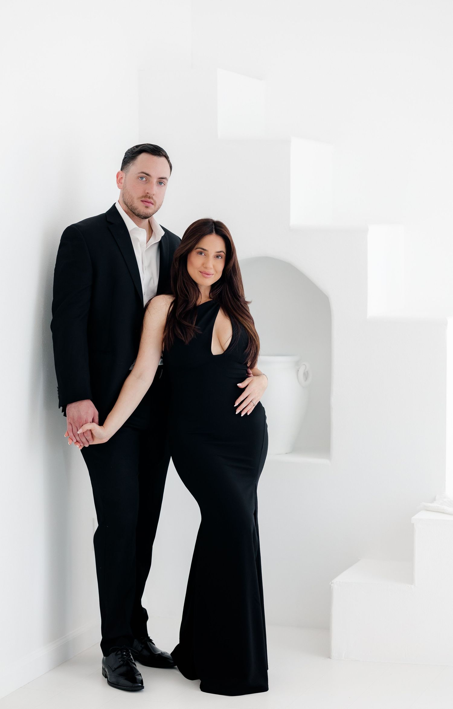 A couple wearing black outfits poses together against a white background in a minimalist maternity photo shoot.