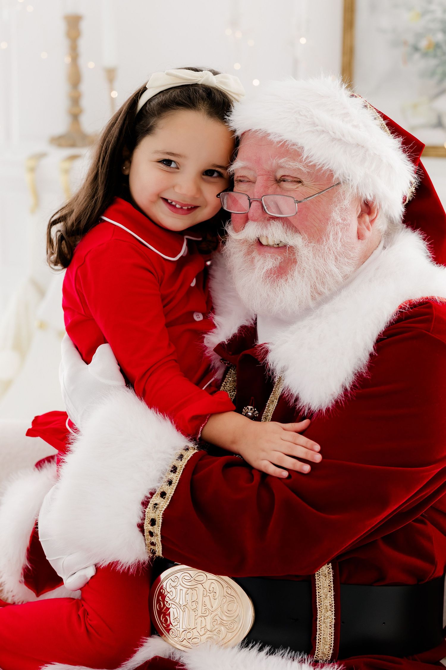 A joyful young girl in red pajamas smiles while sitting on Santa Claus's lap during a Christmas photo session.