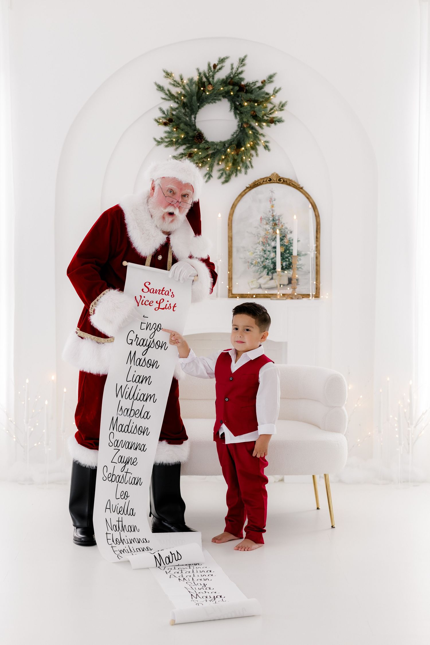 Young boy in red vest meets Santa Claus holding Nice List scroll in white Christmas studio setting.
