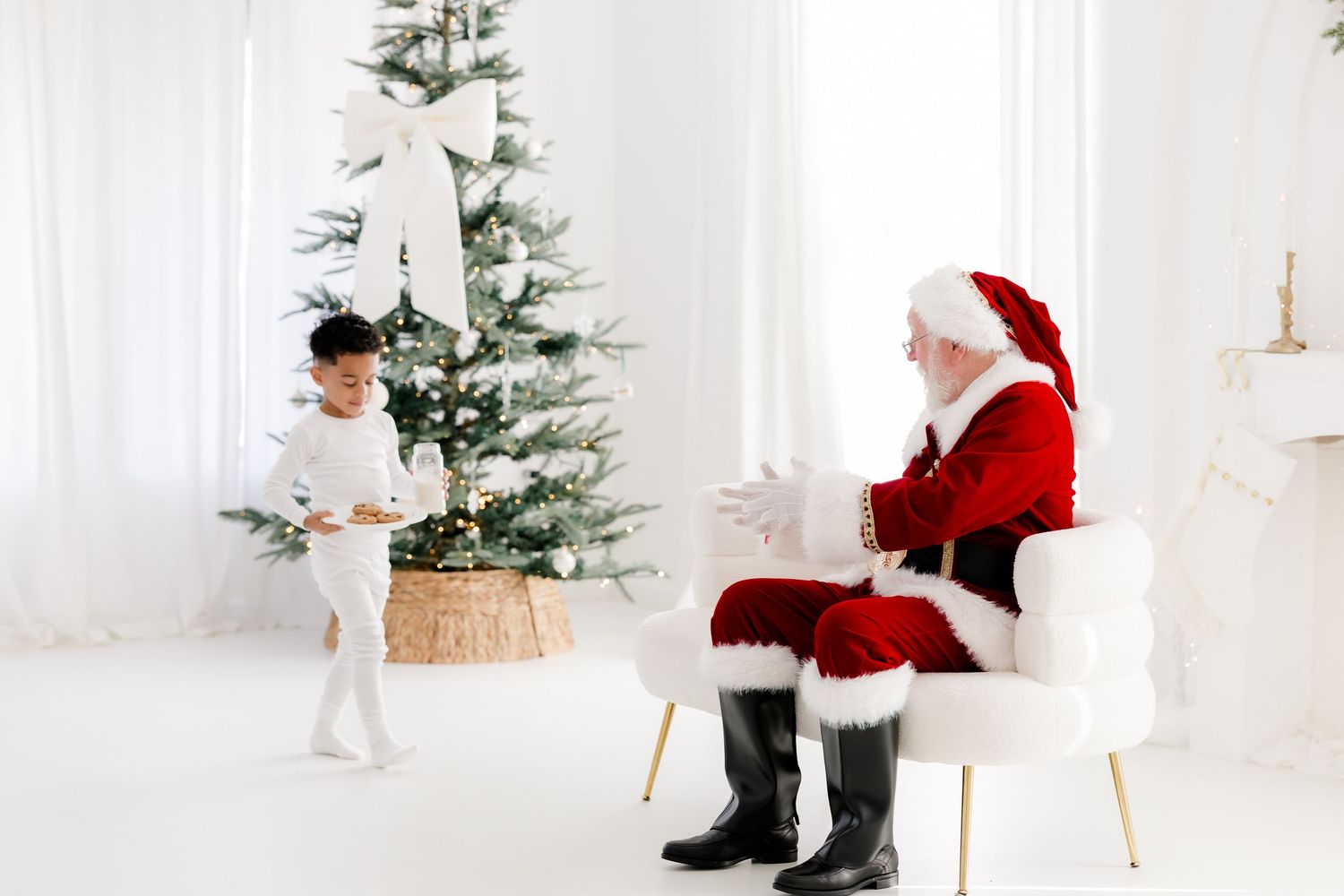 A young child stands before Santa Claus near a decorated white Christmas tree in a bright room.