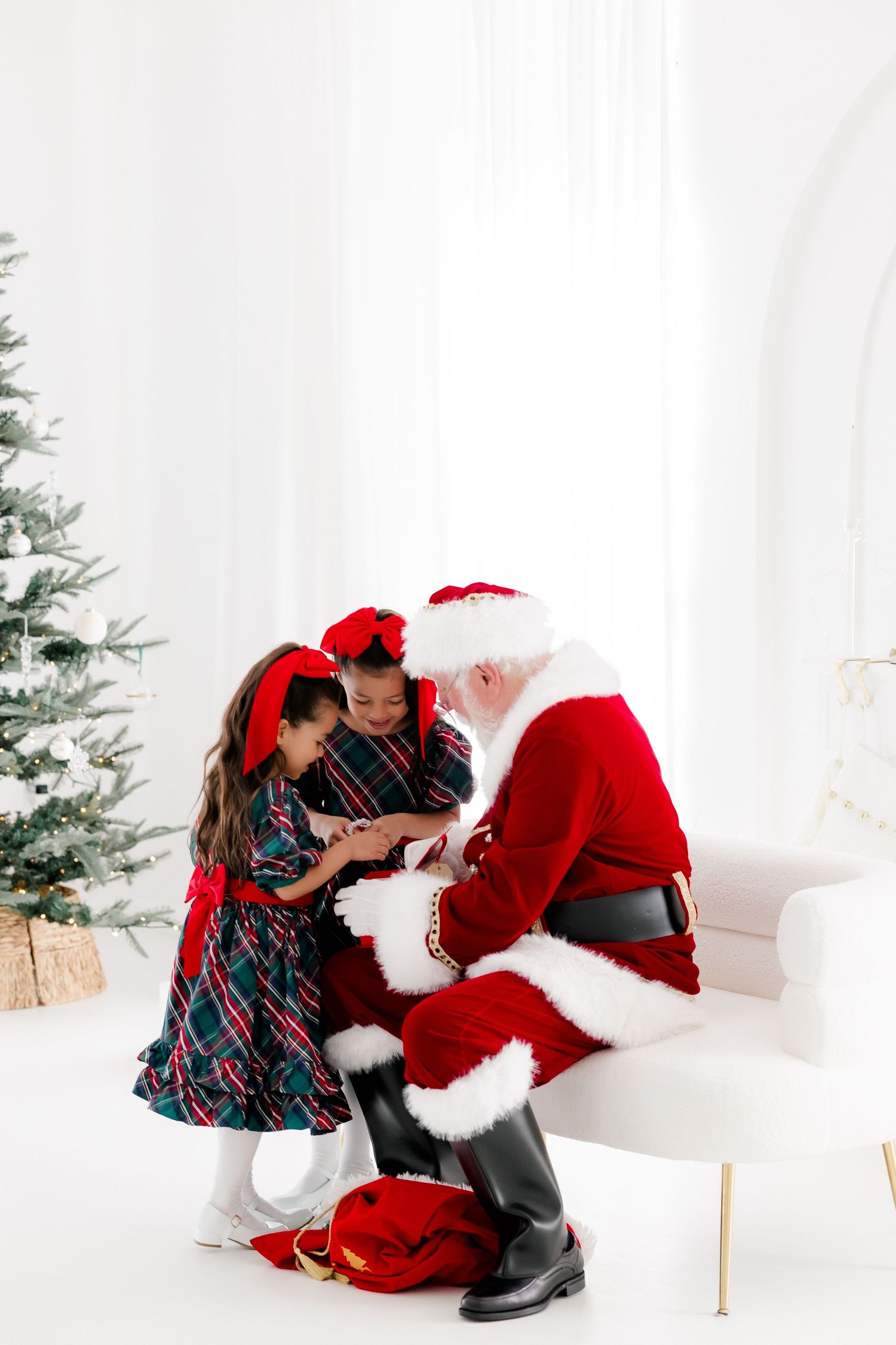 Two children in plaid dresses receive gifts from Santa Claus beside a decorated Christmas tree in a bright white room.