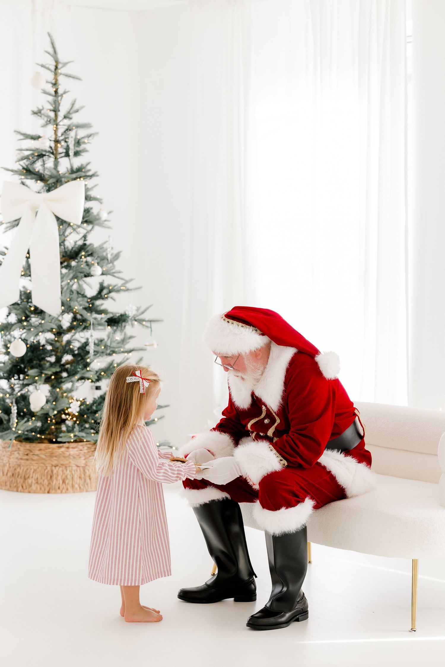 A little girl in a pink dress receives a gift from Santa Claus beside a decorated Christmas tree in a bright white room.