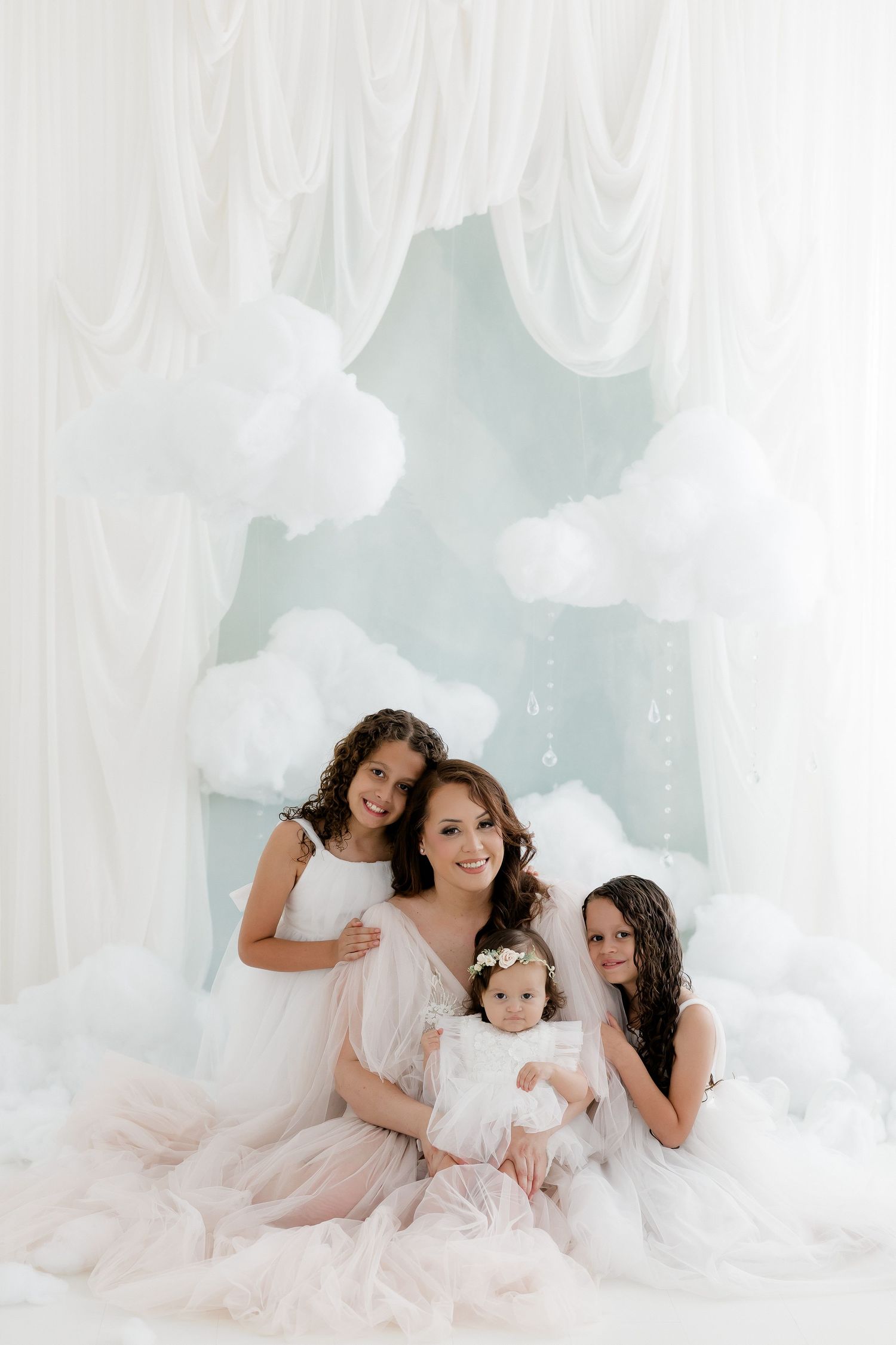 A mother in a pink tulle gown poses with three daughters in white dresses against a dreamy cloud backdrop.