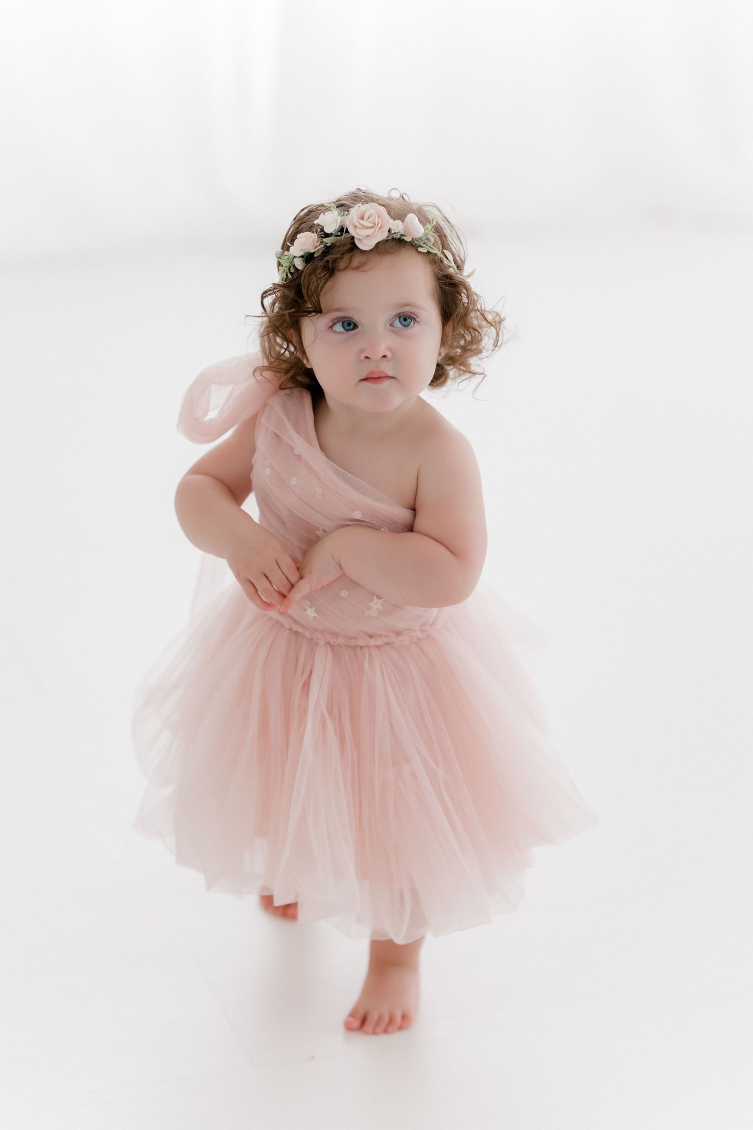 Toddler in pink tulle dress and floral headband poses barefoot against white backdrop in studio portrait.