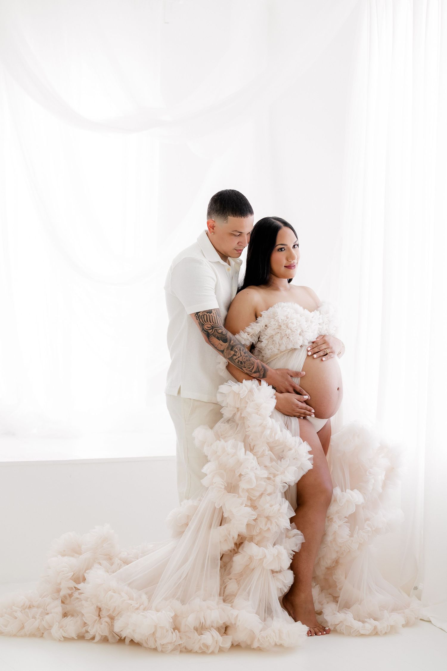 Maternity photoshoot with couple in white featuring flowing tulle dress against light background.