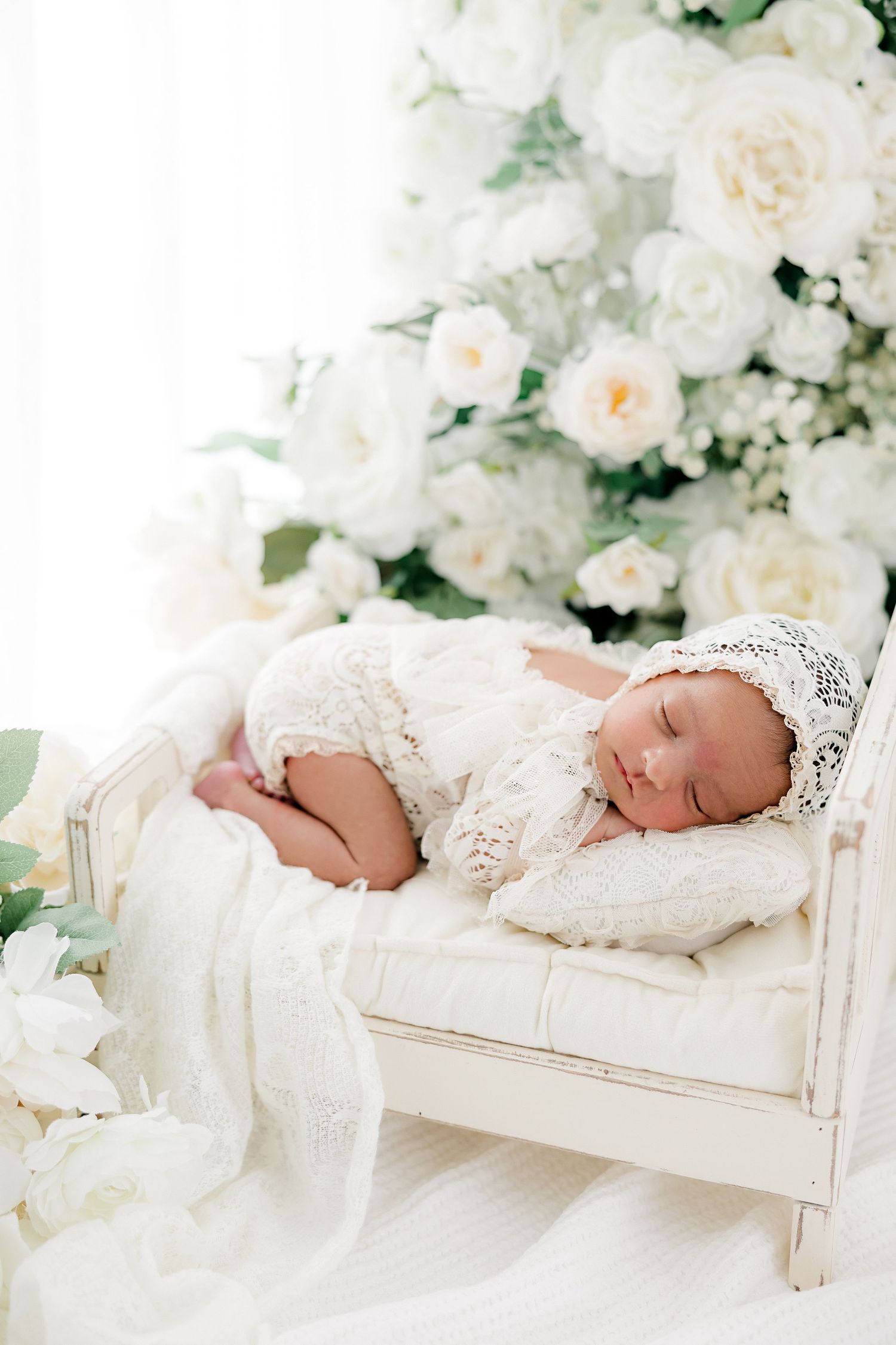 Baby sleeping peacefully on white wooden bed surrounded by cream roses.