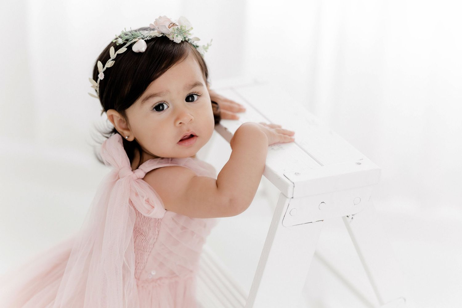 A baby in a delicate pink dress and floral headband poses against a white backdrop in an elegant studio setting.