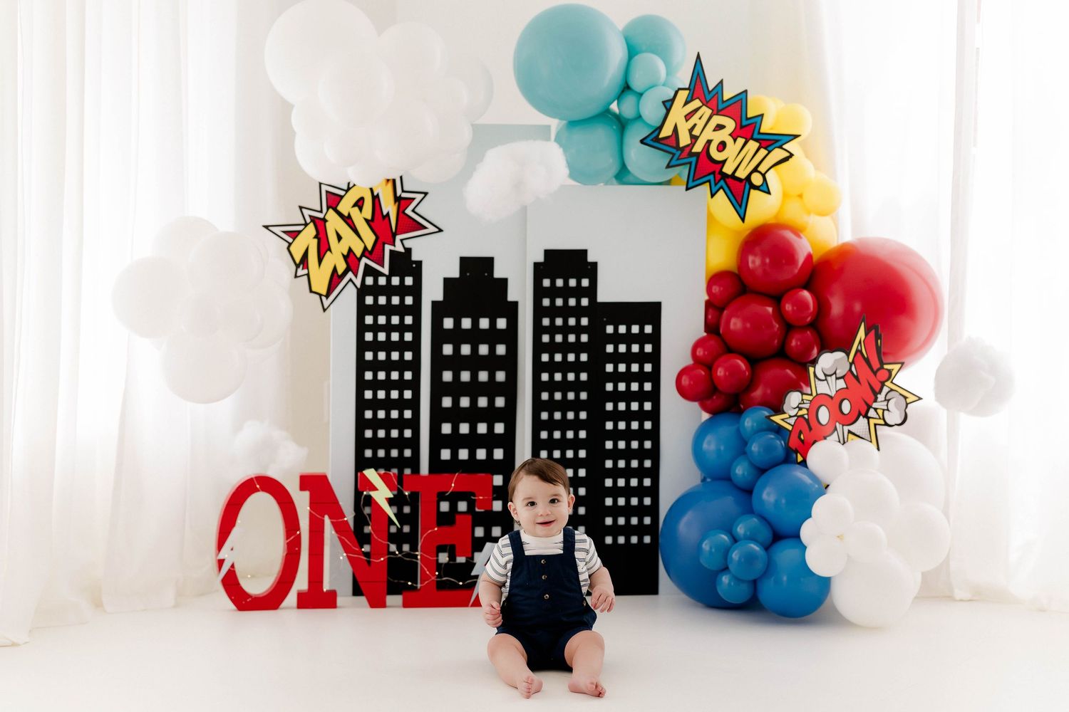 Colorful superhero themed birthday party backdrop with balloon decorations and city skyline silhouettes.