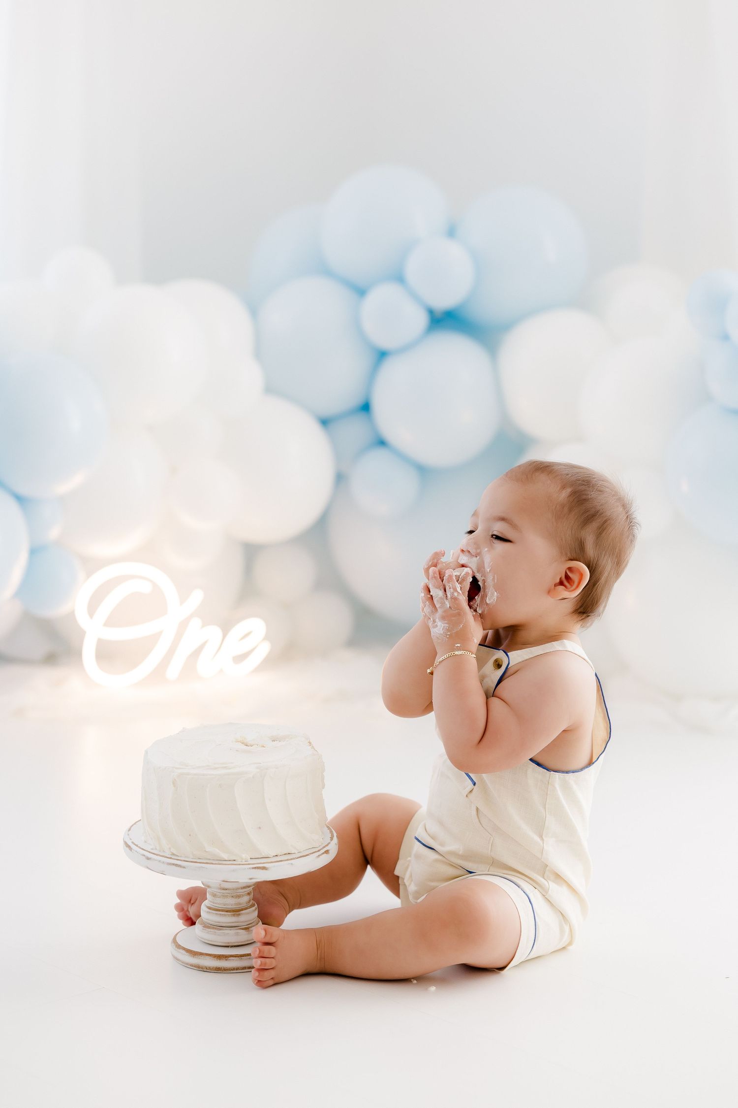 Baby in white outfit sits beside first birthday cake with blue balloon decor and neon 'One' sign in background.