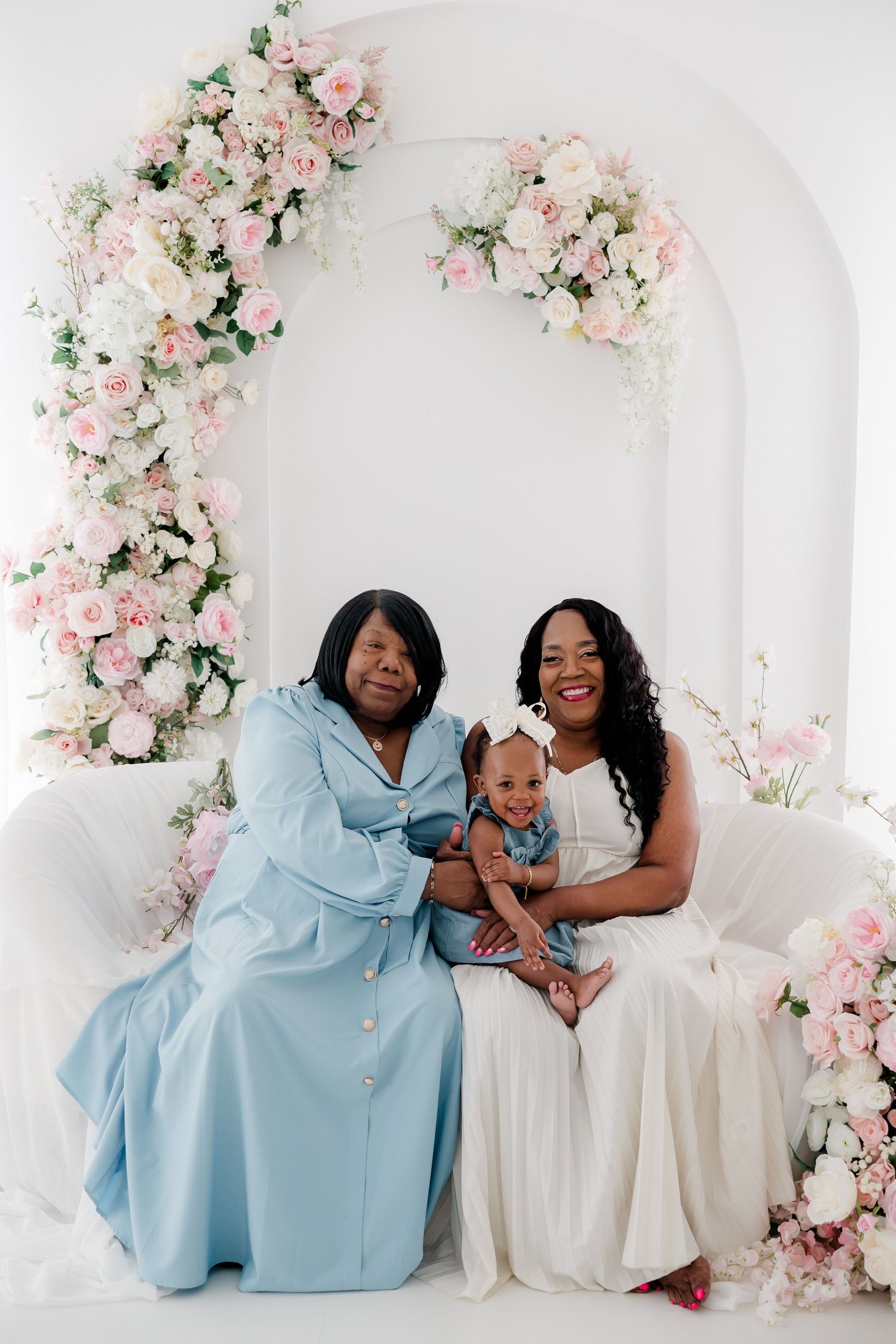 A family of three poses together in a dreamy white studio setting with pink floral decorations and arches.