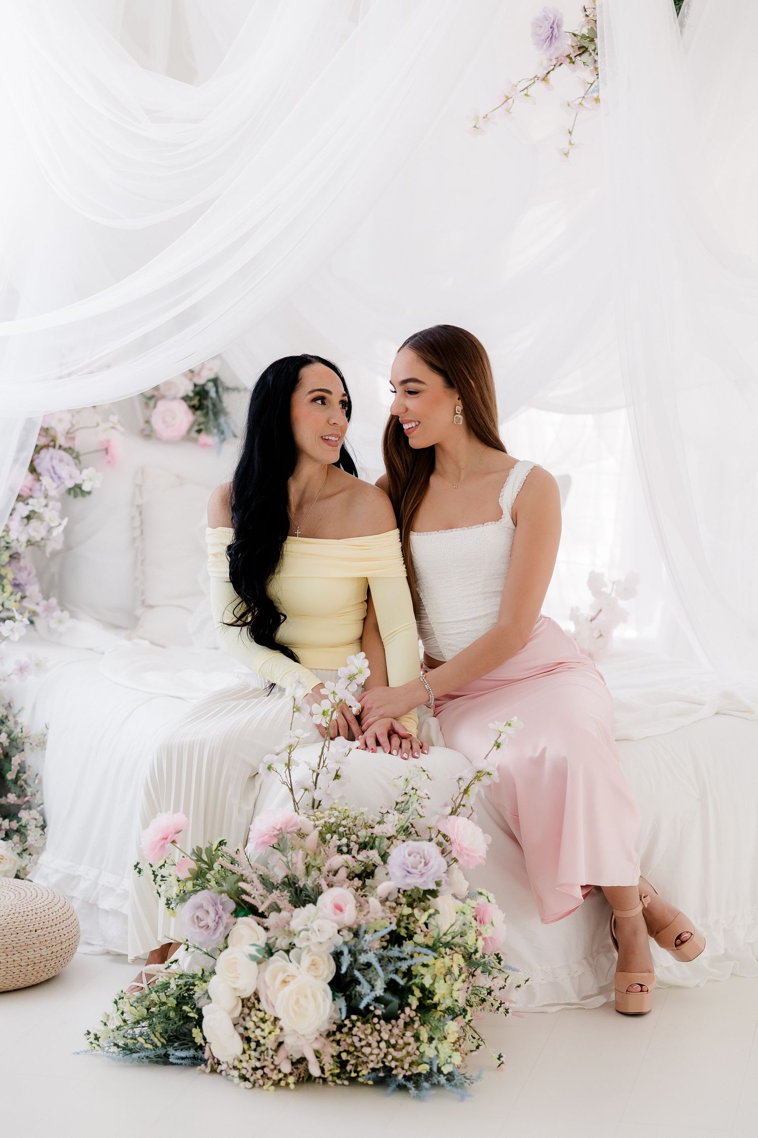 Two women in pastel dresses sit together on white furniture with floral decorations and romantic spring decor.