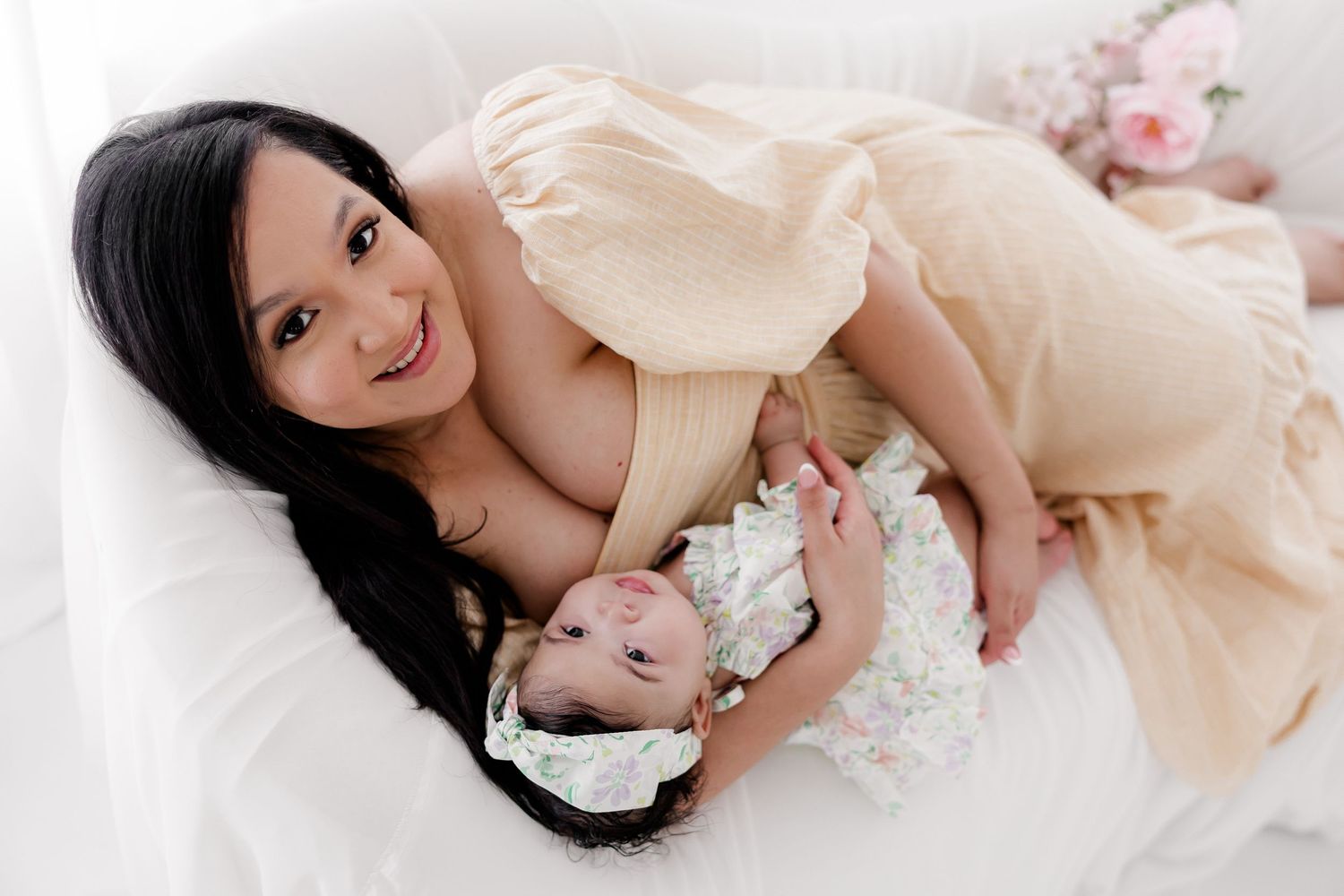 A parent in a cream-colored dress lies on white bedding while embracing a baby wearing a floral outfit.