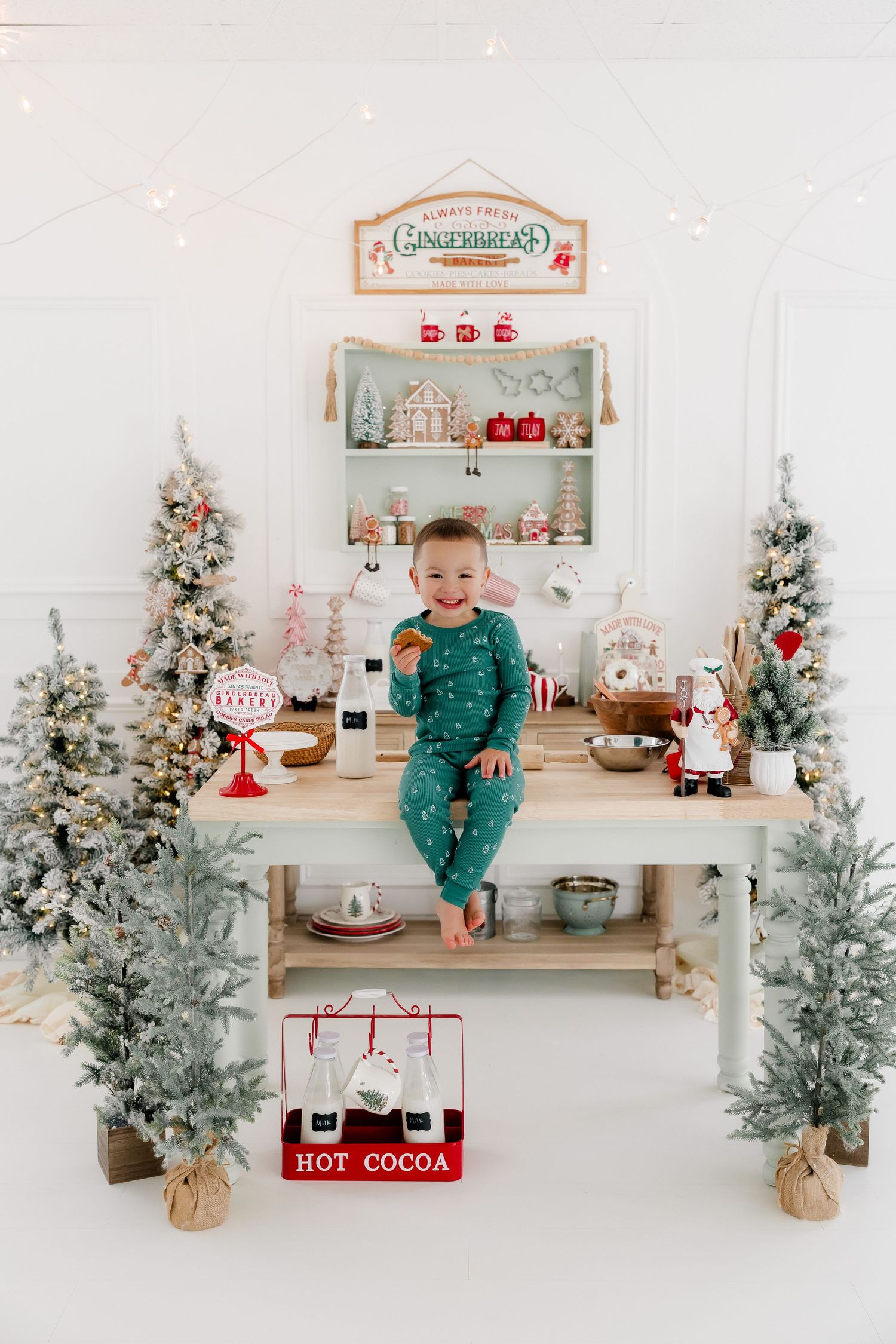 Toddler in green pajamas sits on wooden table surrounded by flocked Christmas trees and holiday gingerbread bakery decor.