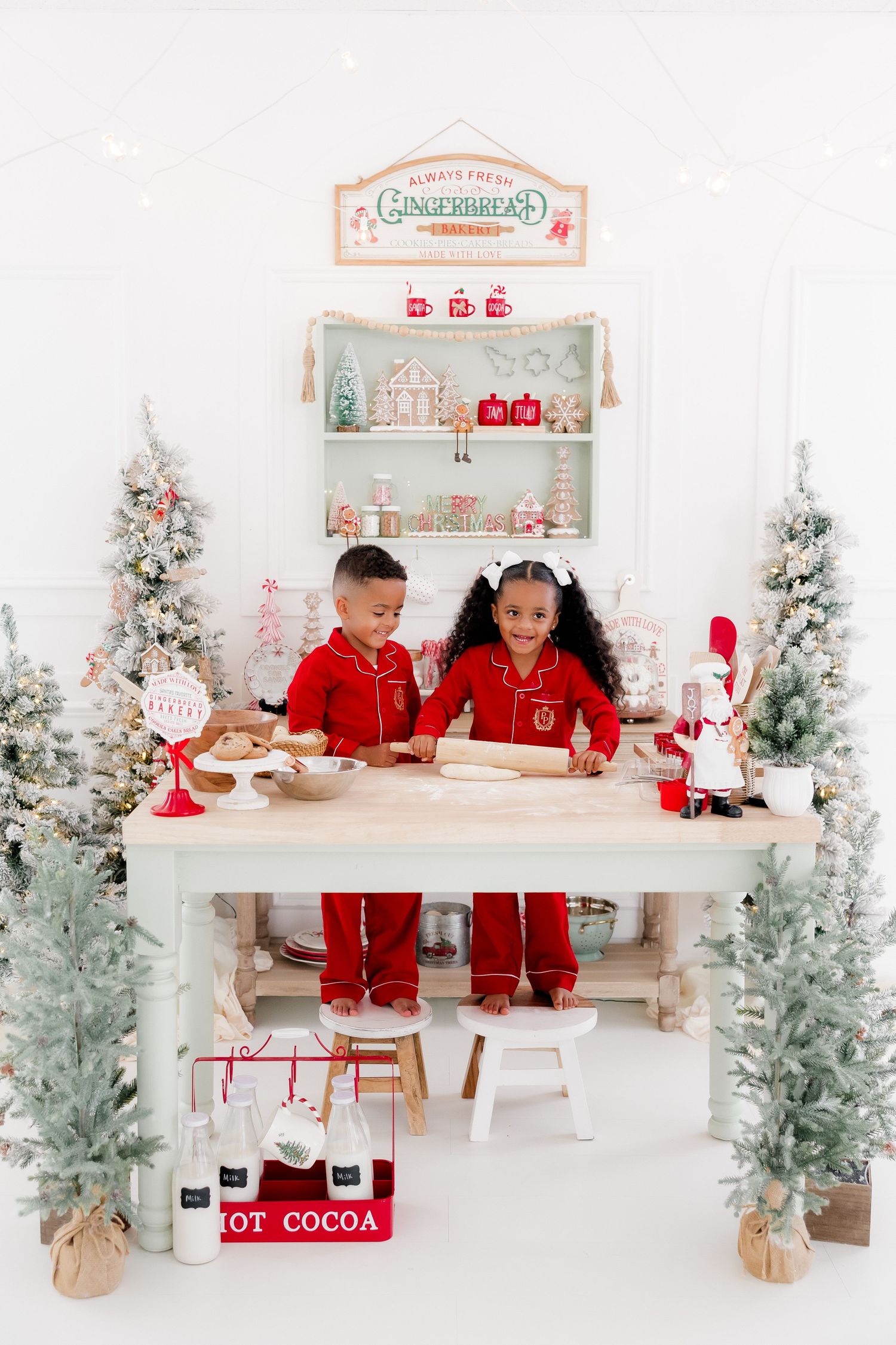 Two children in red pajamas bake Christmas cookies at a festive white table surrounded by snowy trees and holiday decor.