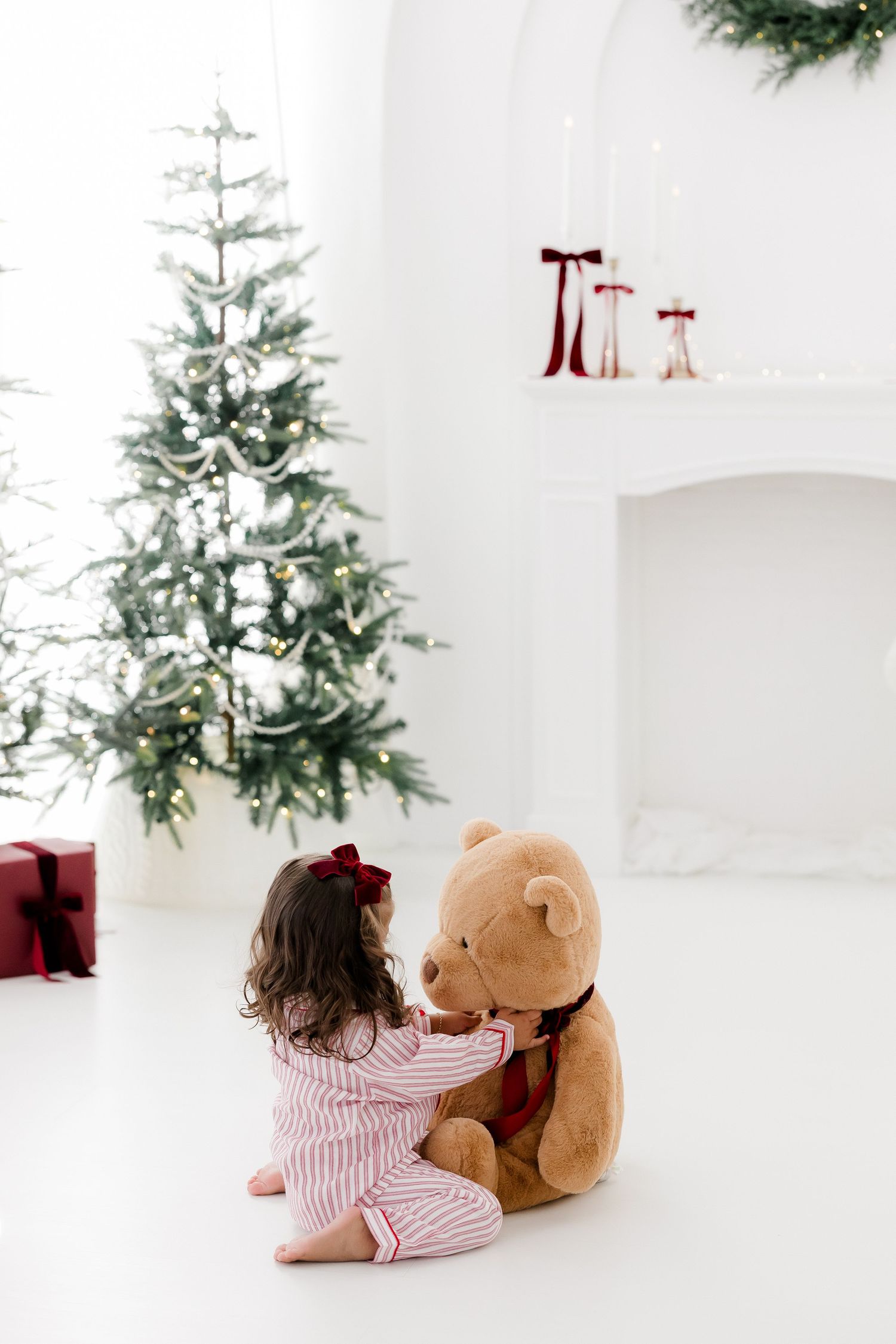 Little girl in pink striped pajamas plays with large teddy bear beside lit Christmas tree in bright white room.