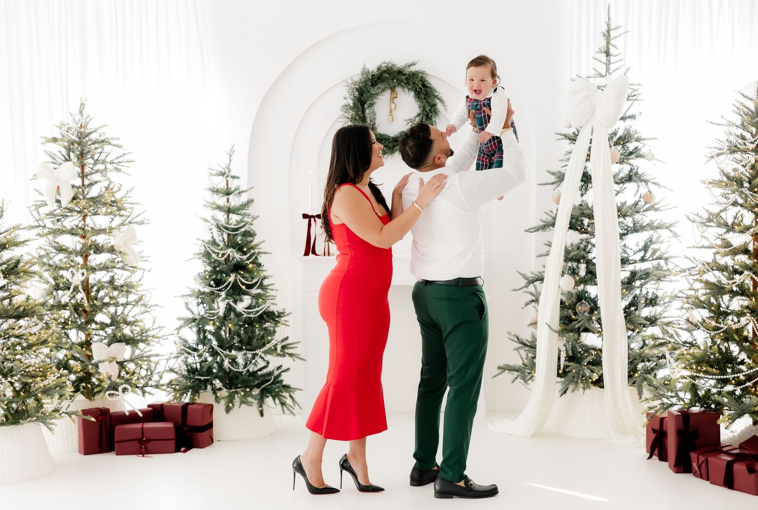 Family Christmas photo with parents holding baby near decorated trees and gifts in white studio.