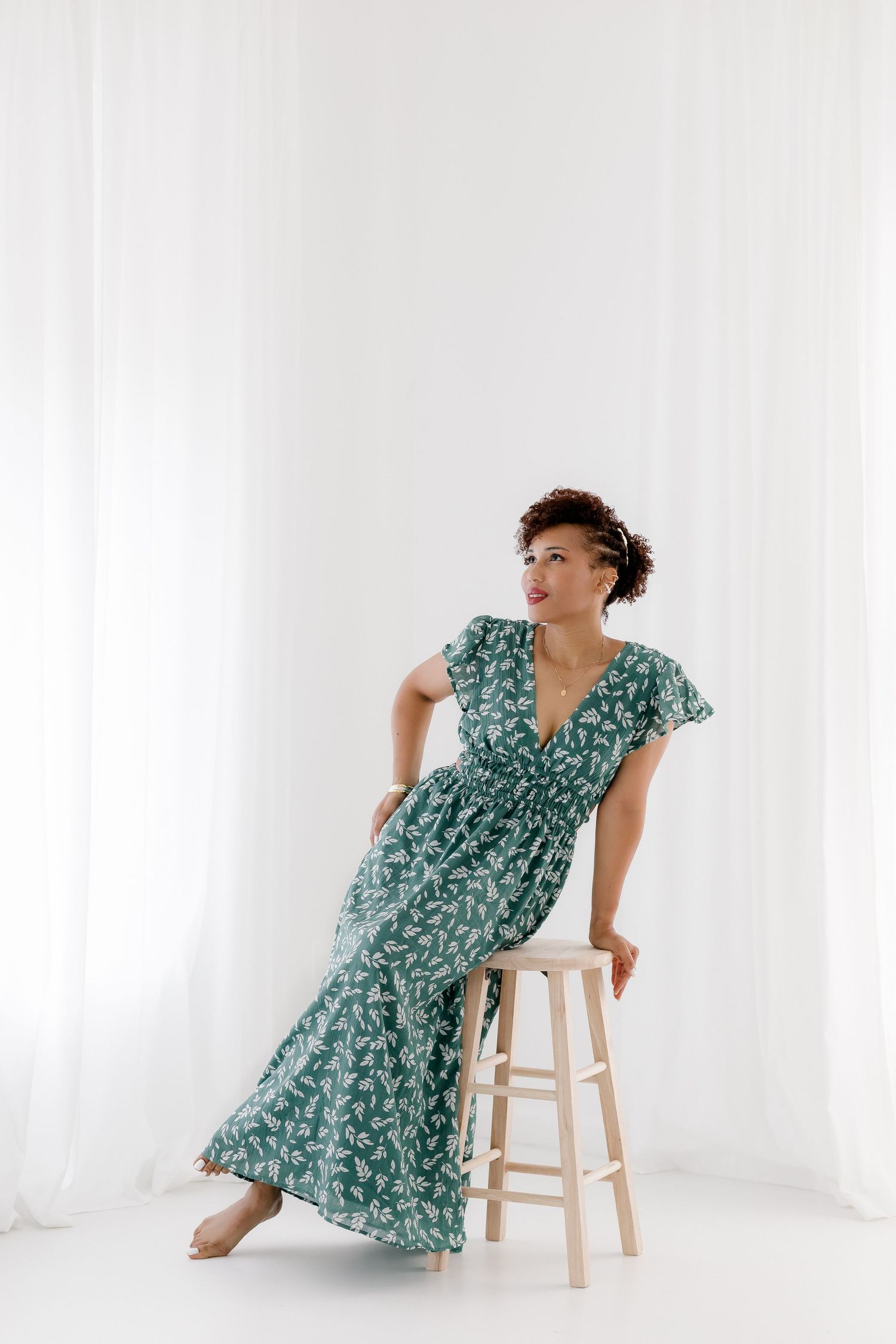 Woman in teal floral dress poses gracefully with wooden stool in bright white studio setting.