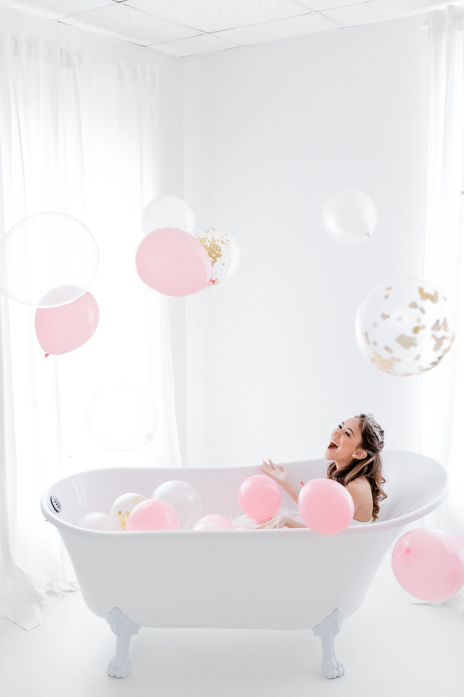 Woman relaxing in vintage clawfoot bathtub with pink and white balloons floating in bright white bathroom.
