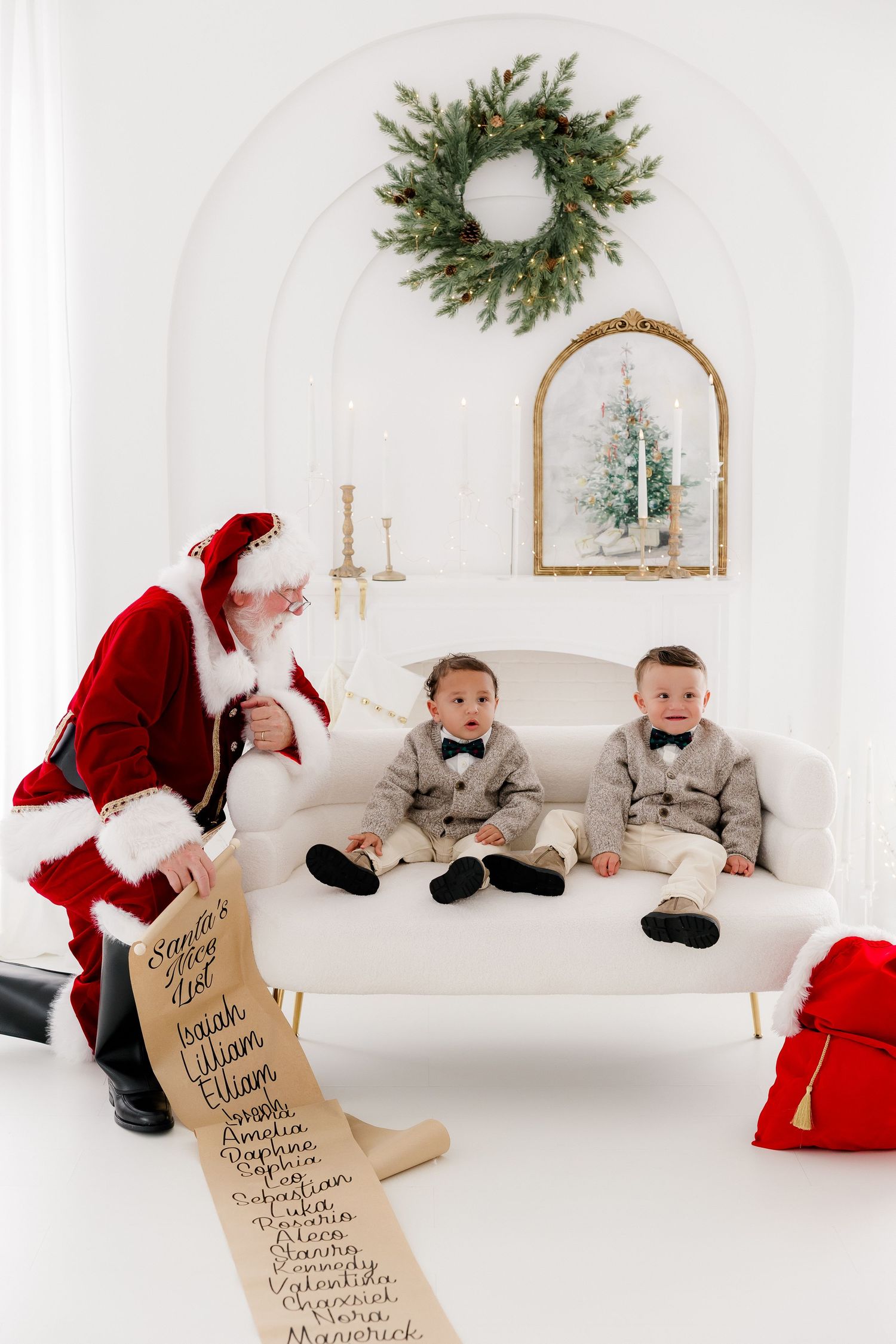 Santa Claus showing his naughty and nice list to two young boys in matching gray suits on a white couch.