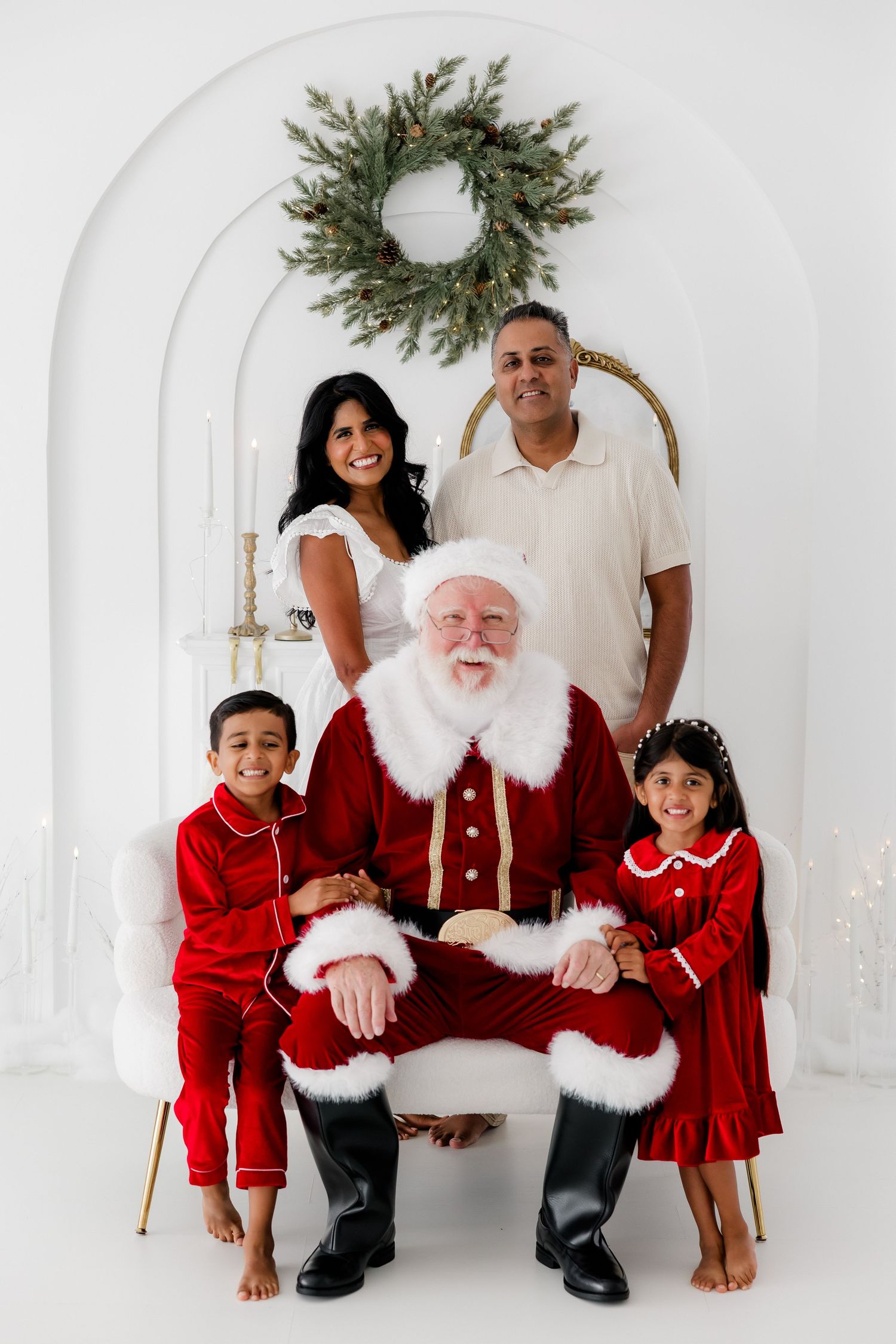 Family posing with Santa Claus in festive red outfits against white arched backdrop with Christmas wreath.