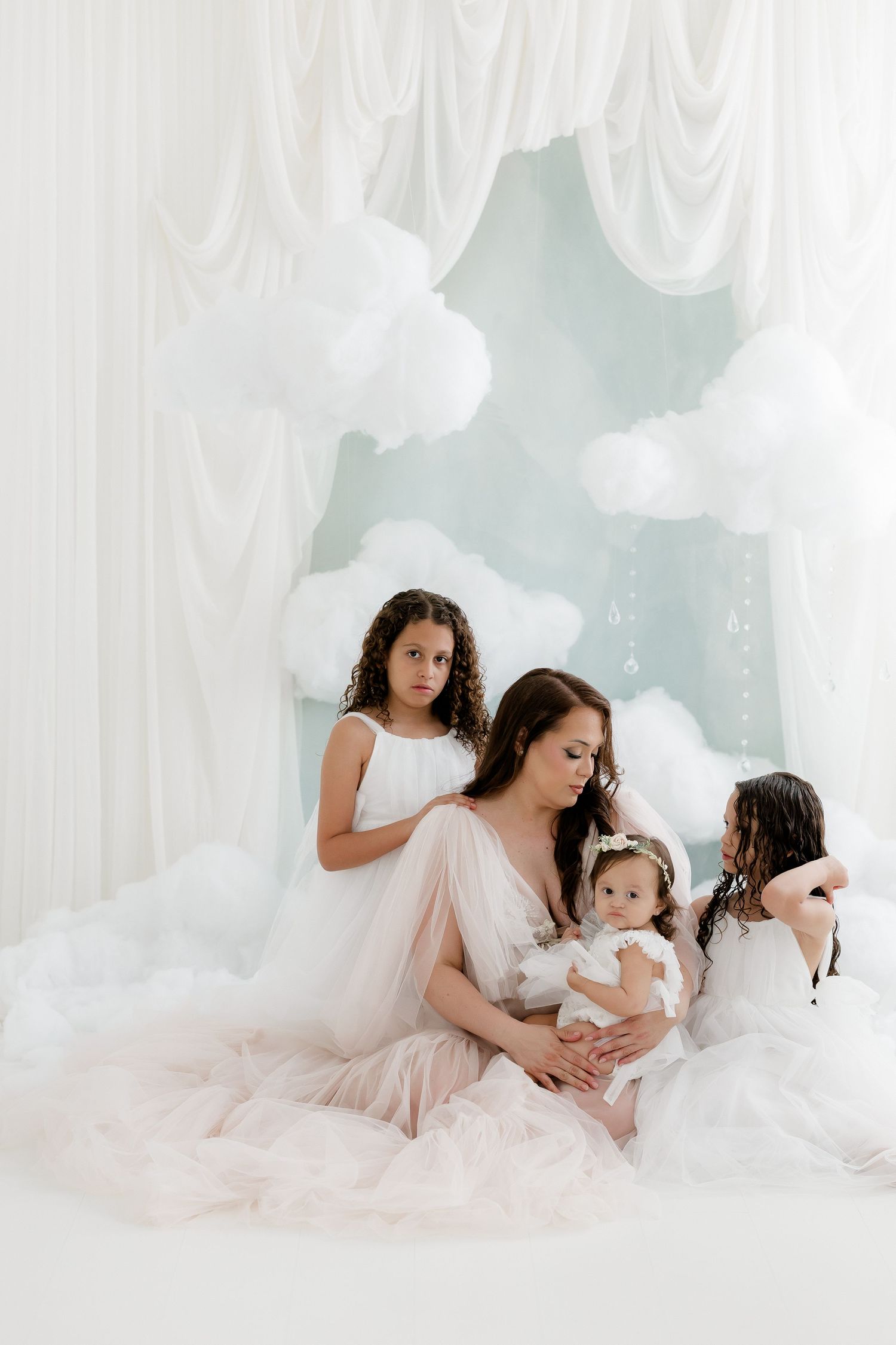 A mother in a white tulle gown sits with three daughters amid cloud decorations in a dreamy white studio setting.