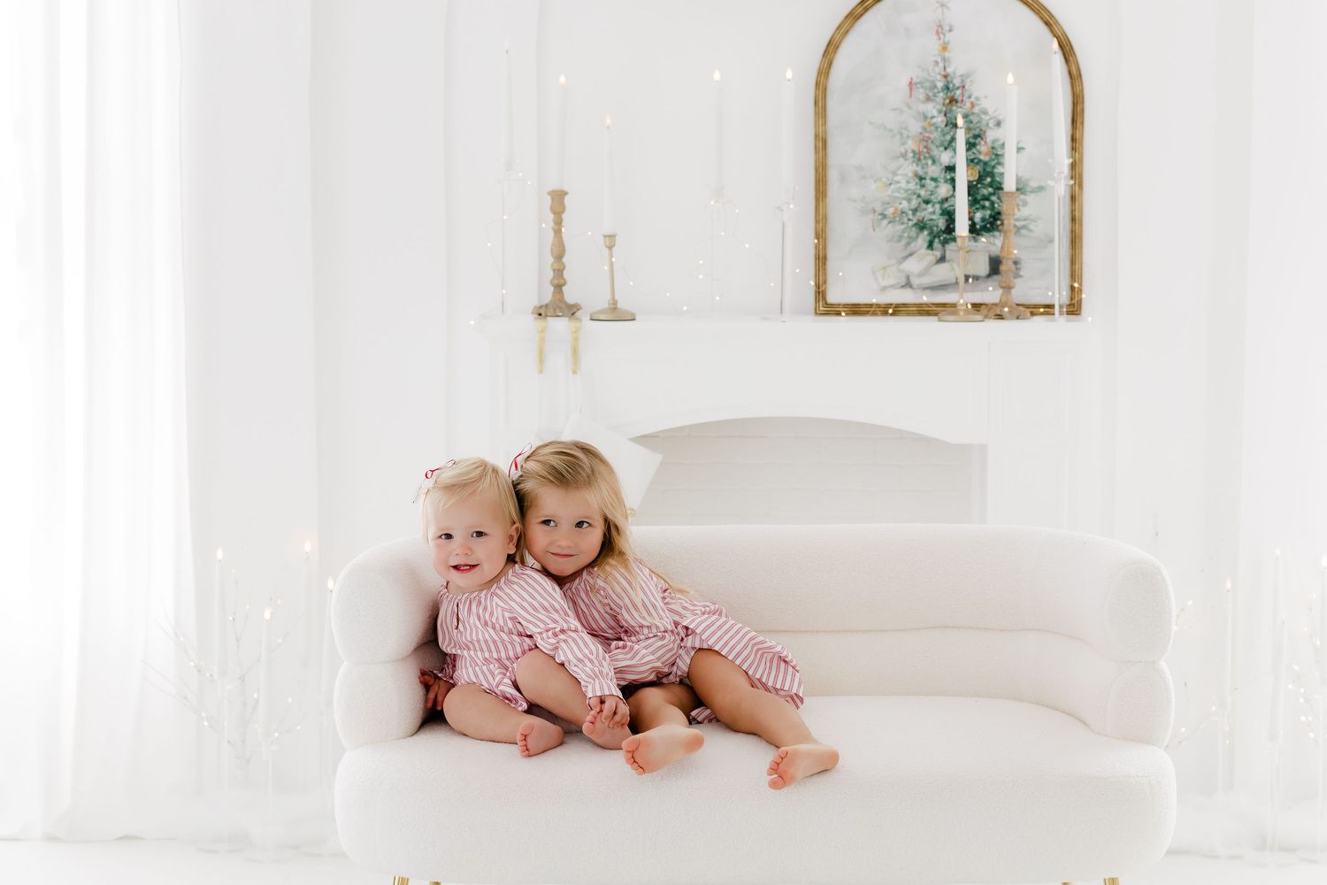 Two young children in matching pink pajamas sitting together on a white sofa near a Christmas fireplace.