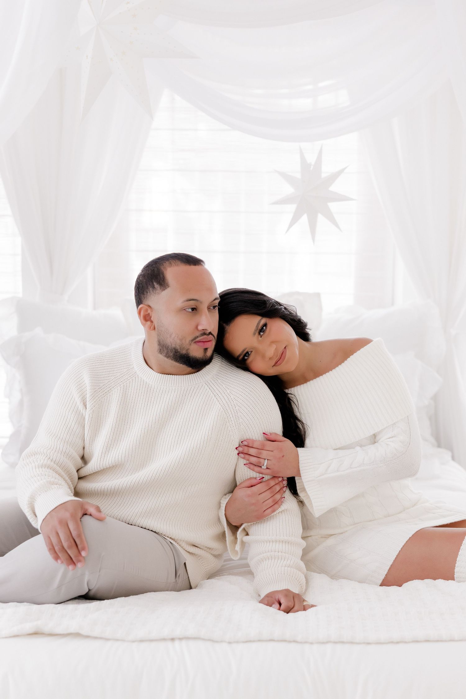 Couple in matching cream sweaters sitting together on a white bed with elegant star window decor.