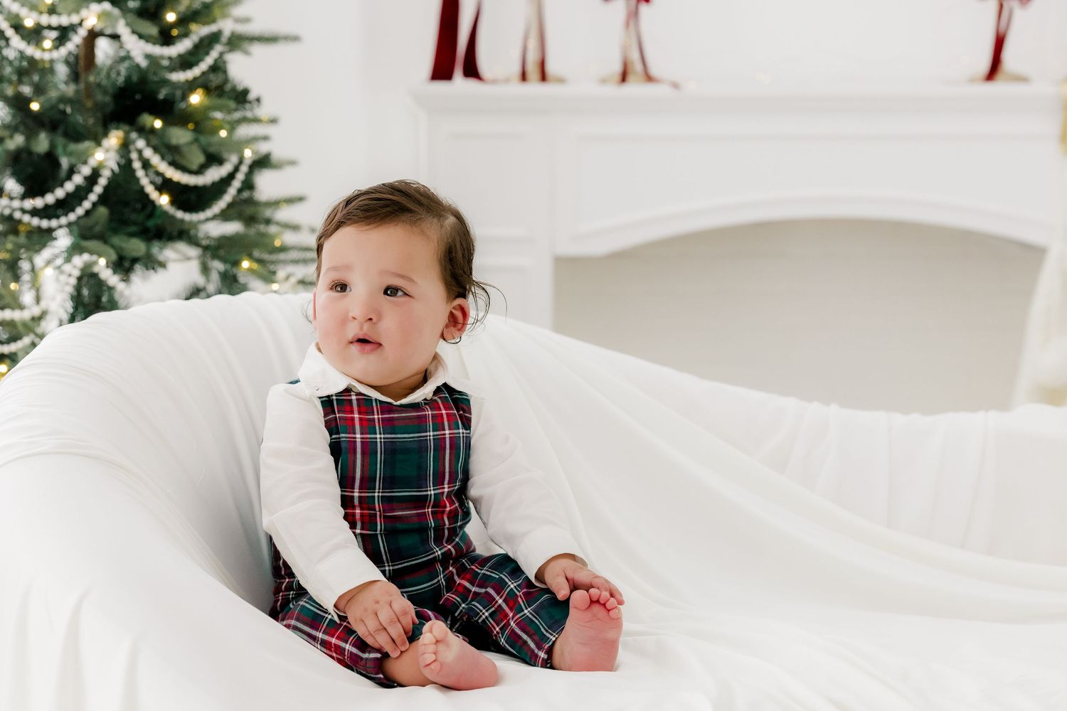 Baby in plaid dress sits on white chair near Christmas tree and fireplace.