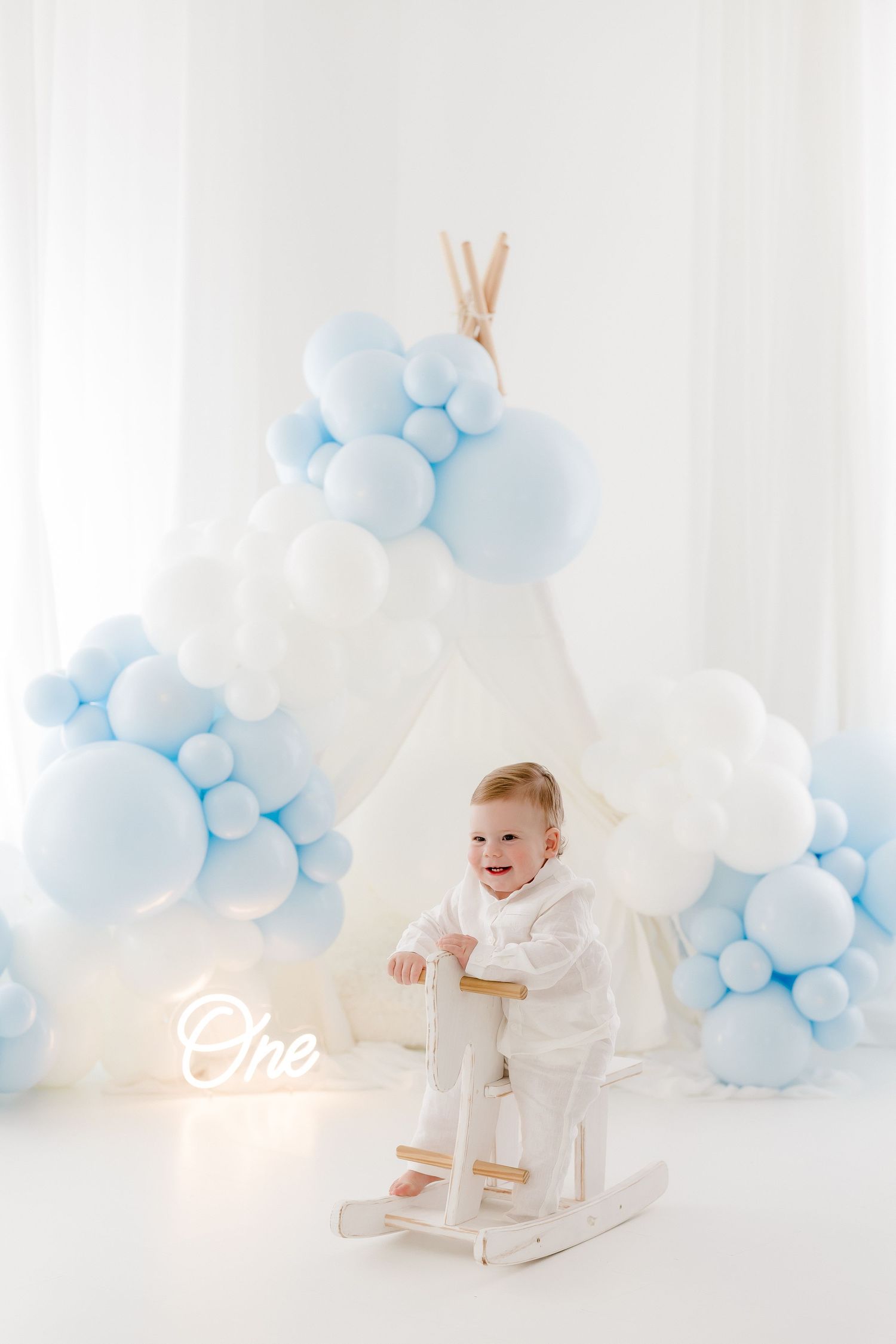 White rocking horse with blue balloon arch decorations and neon sign creates an elegant first birthday photo setup.