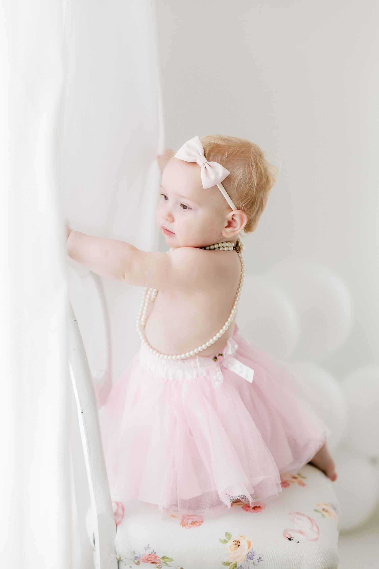 Baby in pink tulle dress and pearls sits gracefully against white backdrop in dreamy portrait session.