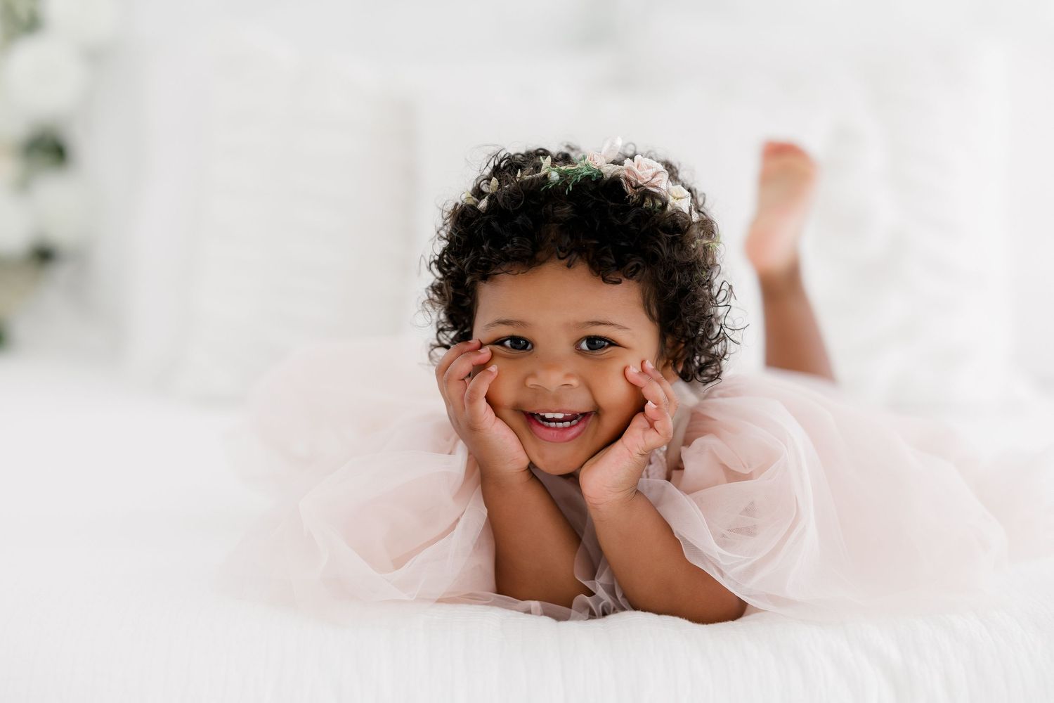 A child in a pink tulle dress lies on a white bed with hands propped under their chin, smiling brightly.