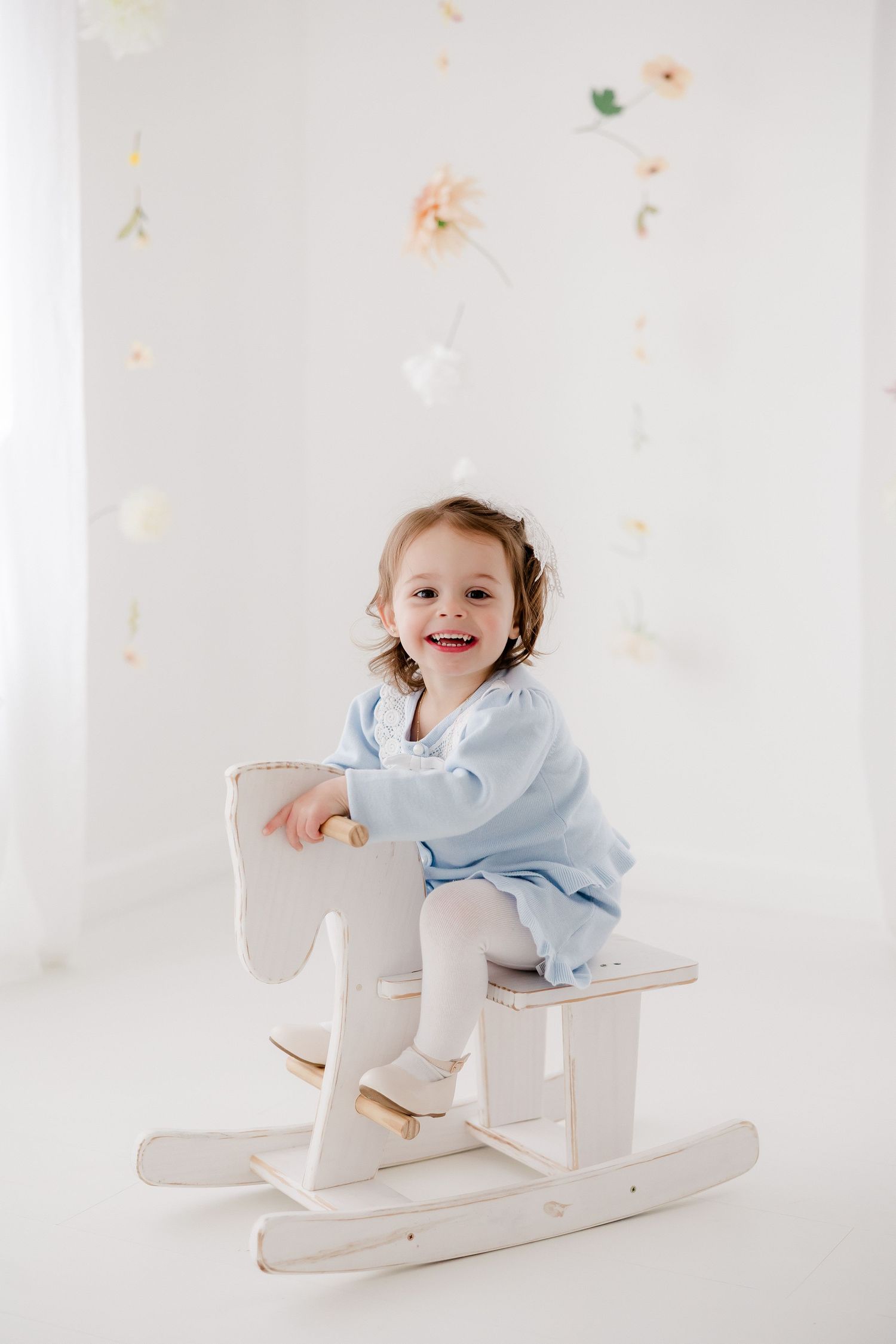 A toddler in a light blue dress sits happily on a white wooden rocking horse against a white background with floral accents.