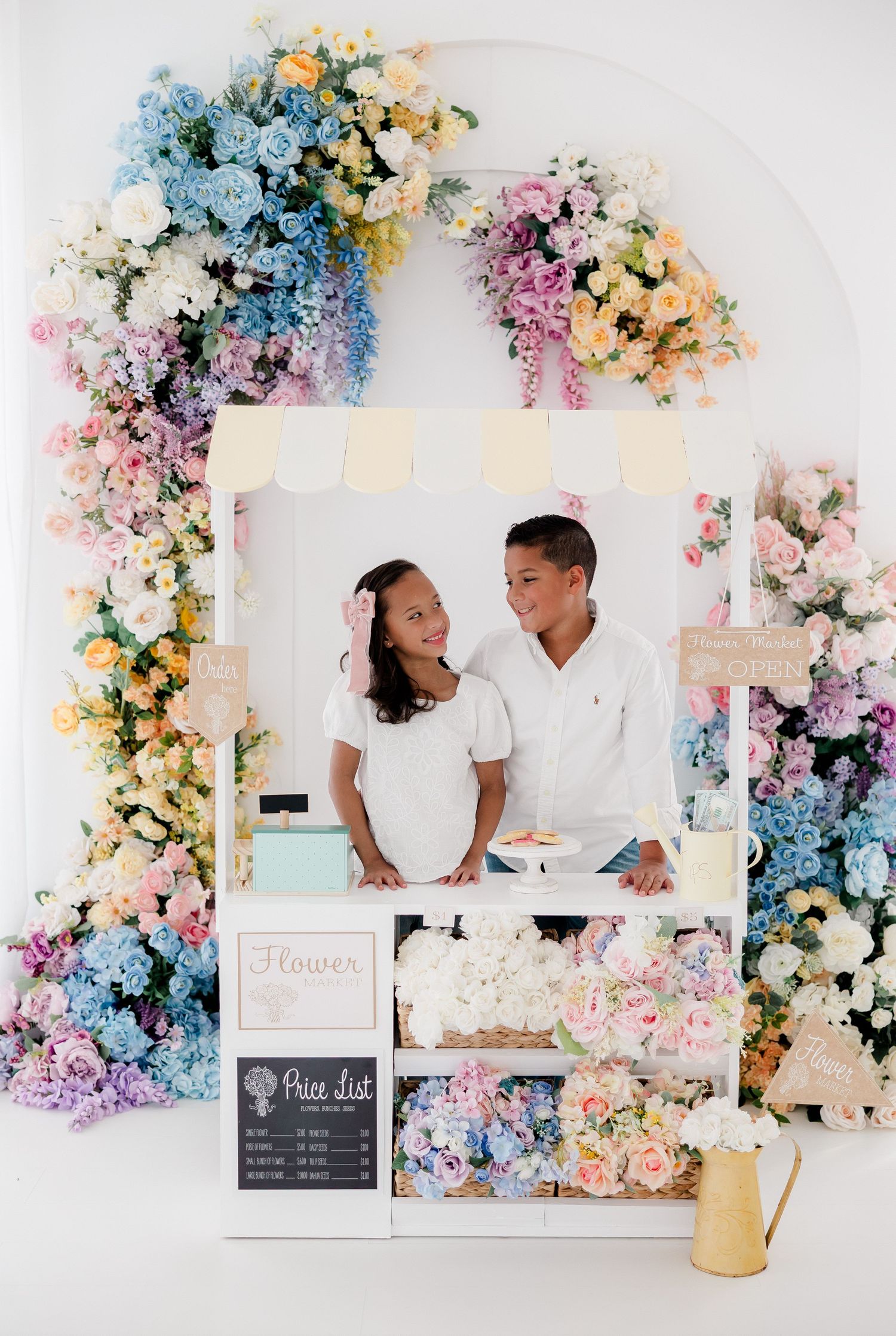 Romantic wedding photo session setup with pastel floral arch featuring blue, pink and yellow blooms against white backdrop.